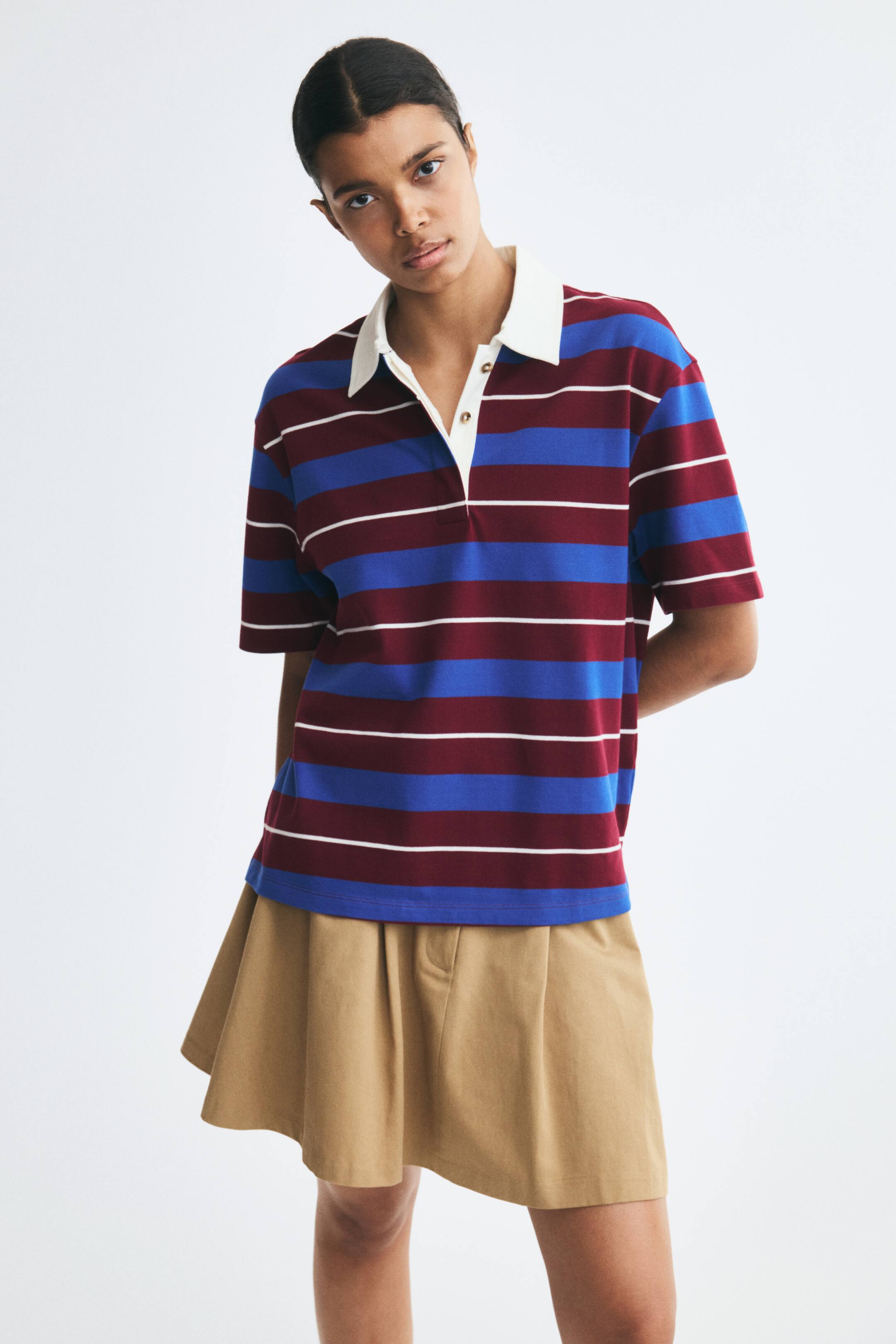 View larger image: The model is wearing a short-sleeved polo shirt featuring horizontal blue, burgundy, and thin white stripes. The shirt is designed with a contrasting white collar and button placket, and is styled with a pleated beige skirt.