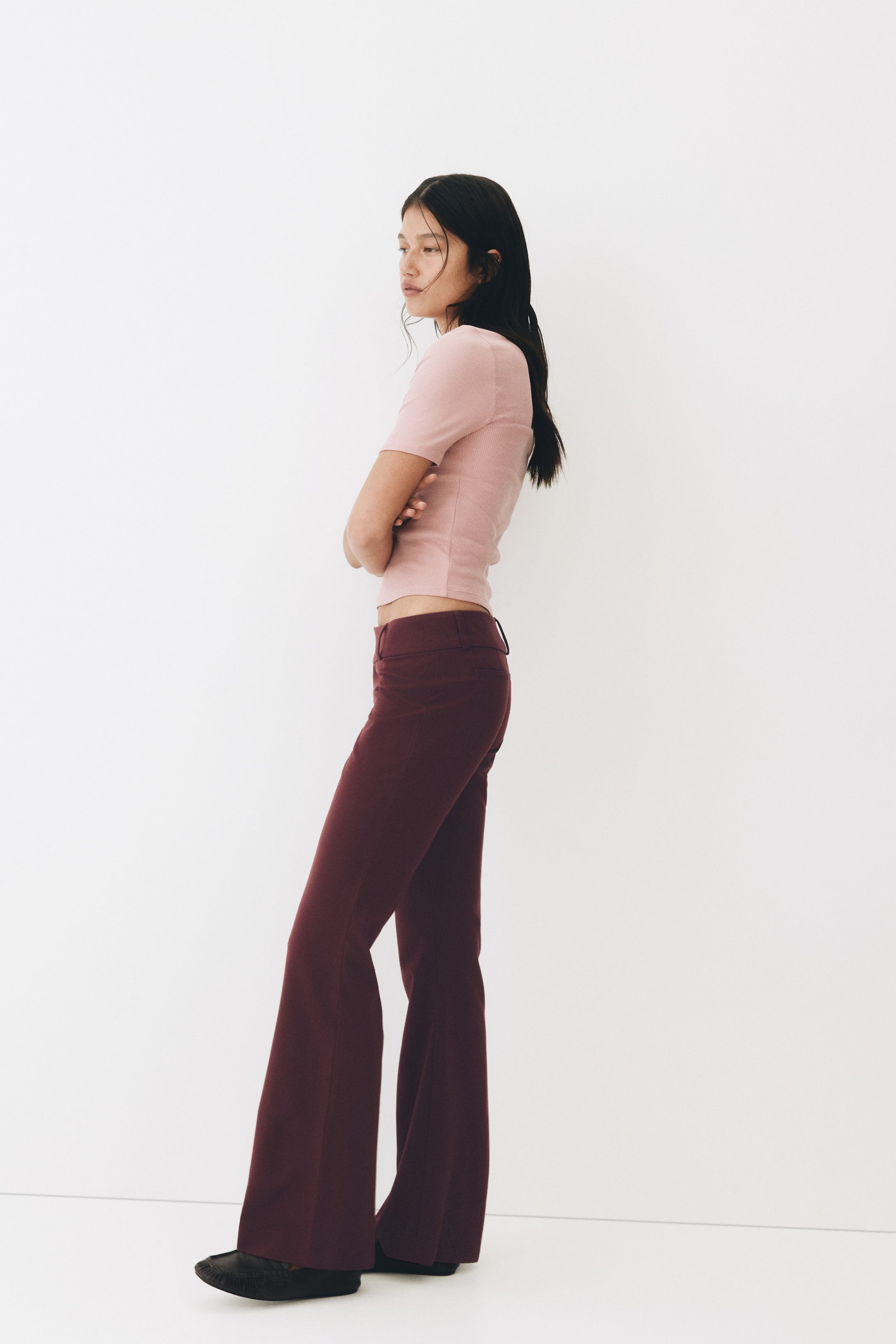 View larger image: The model is wearing burgundy fitted, tailored trousers with a low waist and flared legs.