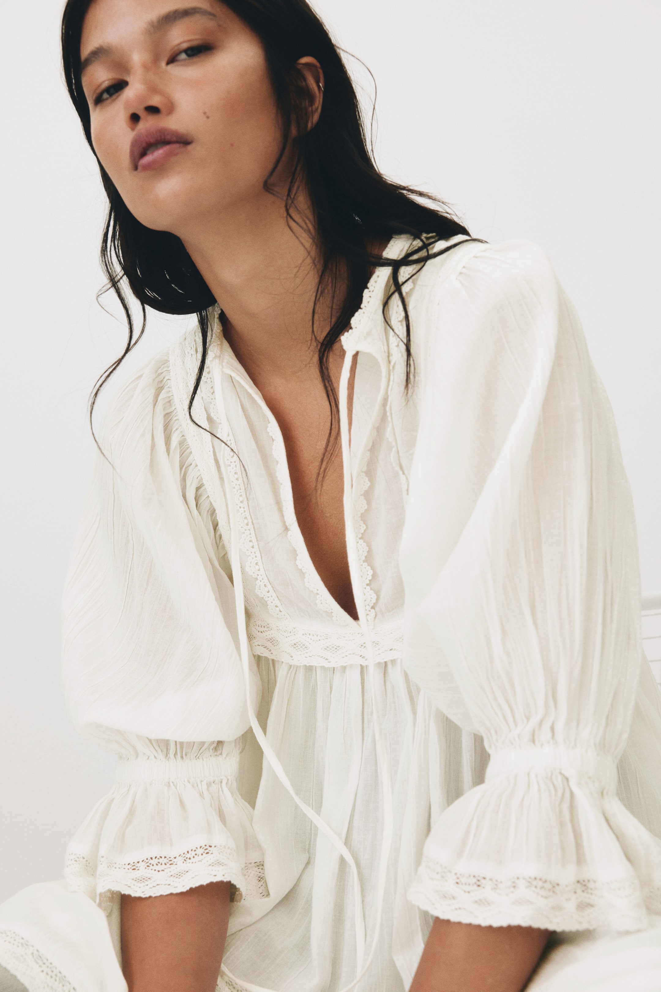 Vis større bilde: Den hvite kjolen har en dyp V-utringning med knytebånd og blondekant langs stolpen og empirelivet. De voluminøse puffermene samles i smockede, rysjede mansjetter som også viser frem blonder.