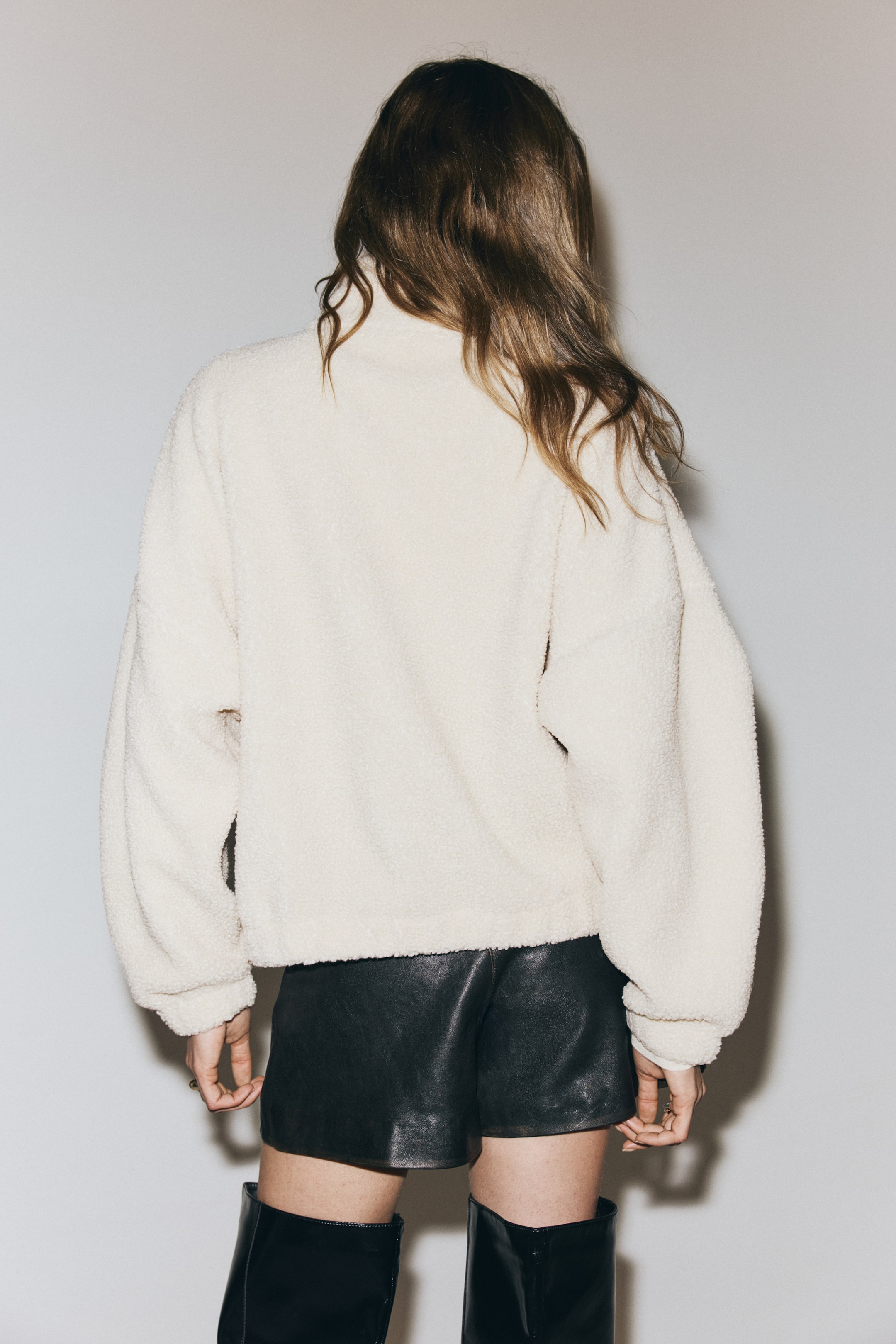 Ingrandisci l'immagine: Un pullover in pile con texture crema è indossato in vista posteriore, caratterizzato da un colletto alto, maniche lunghe e rilassate con polsini a fascia e un orlo che si ferma all’altezza dei fianchi sopra pantaloncini neri.