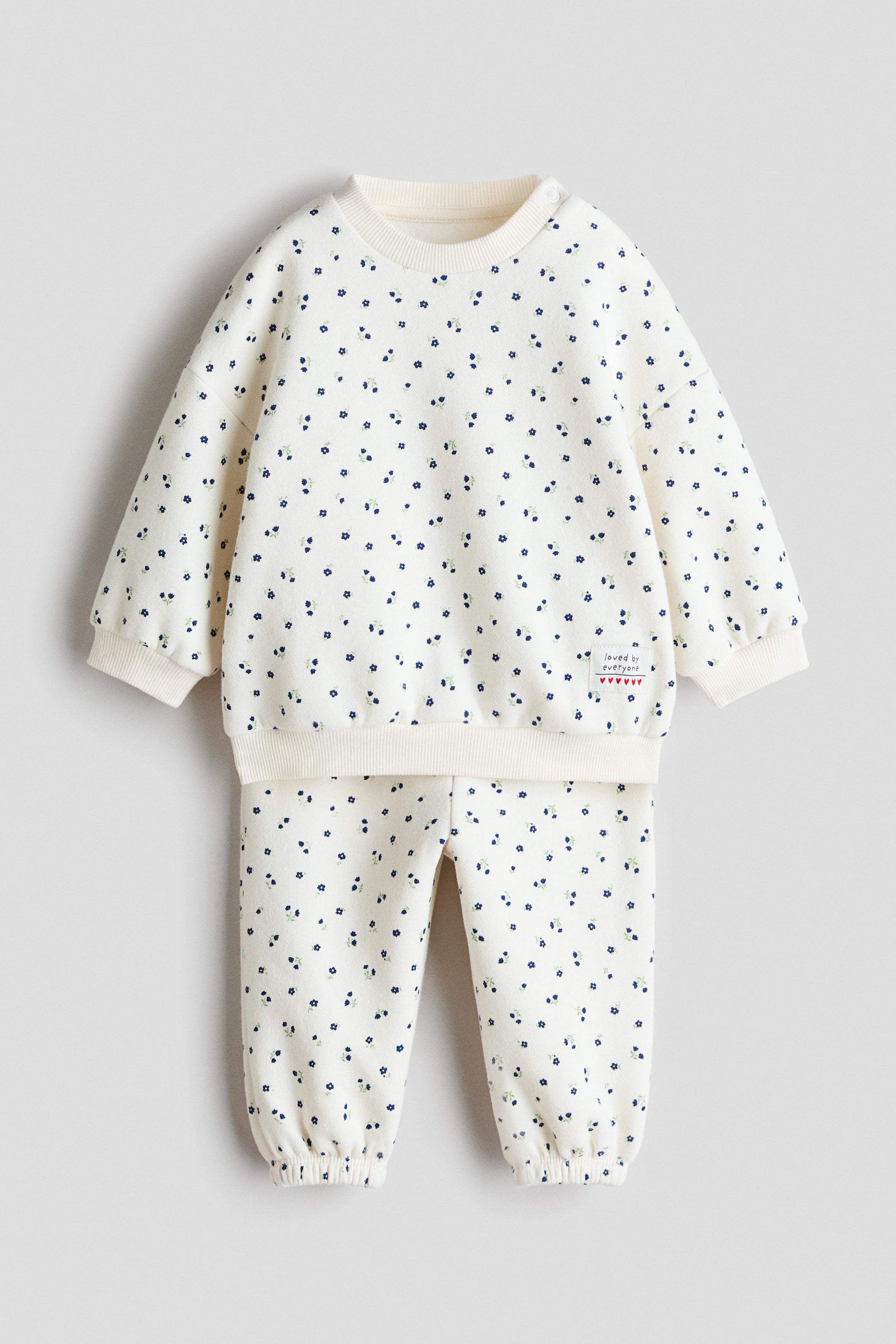 View larger image: A baby's two-piece outfit is shown from the front, featuring a cream-coloured sweatshirt and matching trousers, both adorned with a scattered small blue floral print. The sweatshirt has a ribbed crew neck, ribbed cuffs, and a ribbed hem, with a small white fabric tag on its front right side, while the trousers feature elasticated ankles.