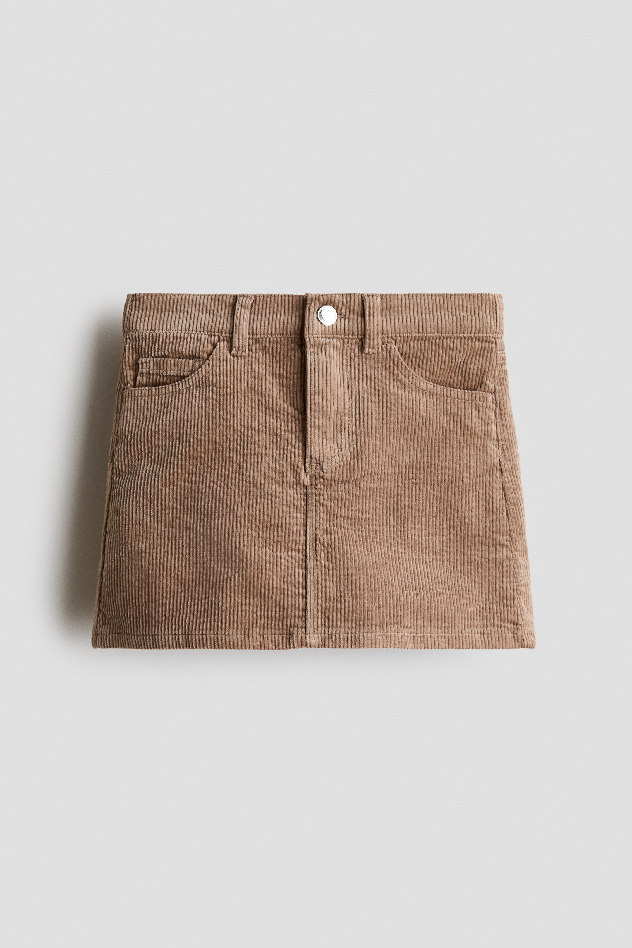 View larger image: A brown corduroy skirt is shown from the front, featuring a waistband with belt loops, a silver button closure, and two curved front pockets, including a smaller coin pocket on the right.