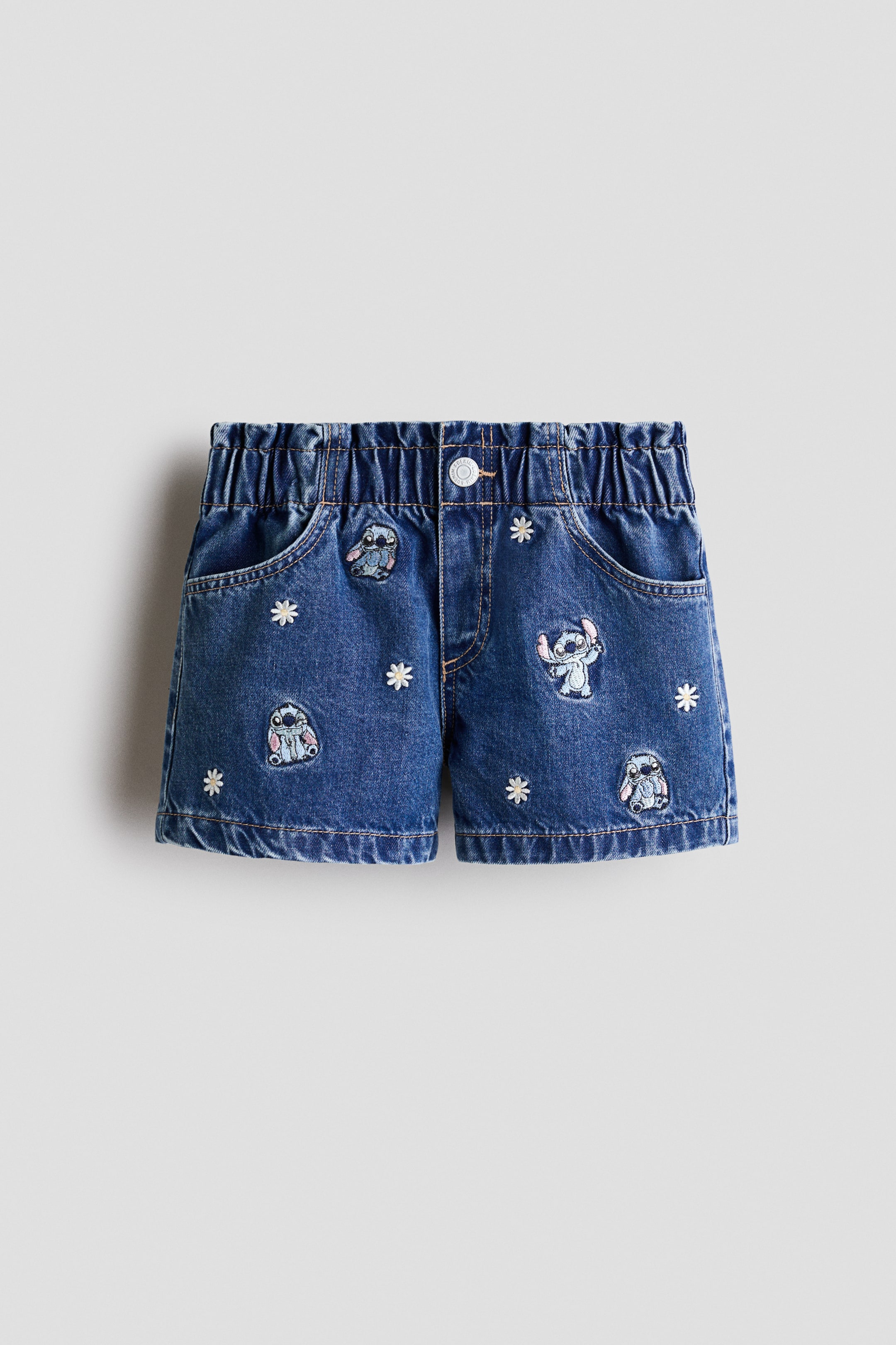 View larger image: Denim shorts are showcasing the front, featuring an elastic waistband with a single button closure, two front pockets, and various embroidered Stitch character patches and white daisy flowers scattered across the medium-dark wash denim.