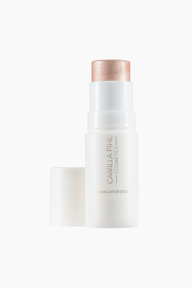 En hvid highlighter-stick fra Camilla Pihl Cosmetics står oprejst og viser sit skinnende fersken-guldfarvede produkt, der er drejet op fra toppen. Dens hvide hætte ligger vandret ved siden af stiftens bund. - 1