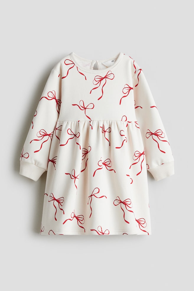 A cream-colored dress with an all-over red bow print is being shown facing FRONT. It is featuring a round neckline with a keyhole opening and button at the back, dropped shoulders, long puff-sleeves with ribbed cuffs, and gathers at the waist. - 1