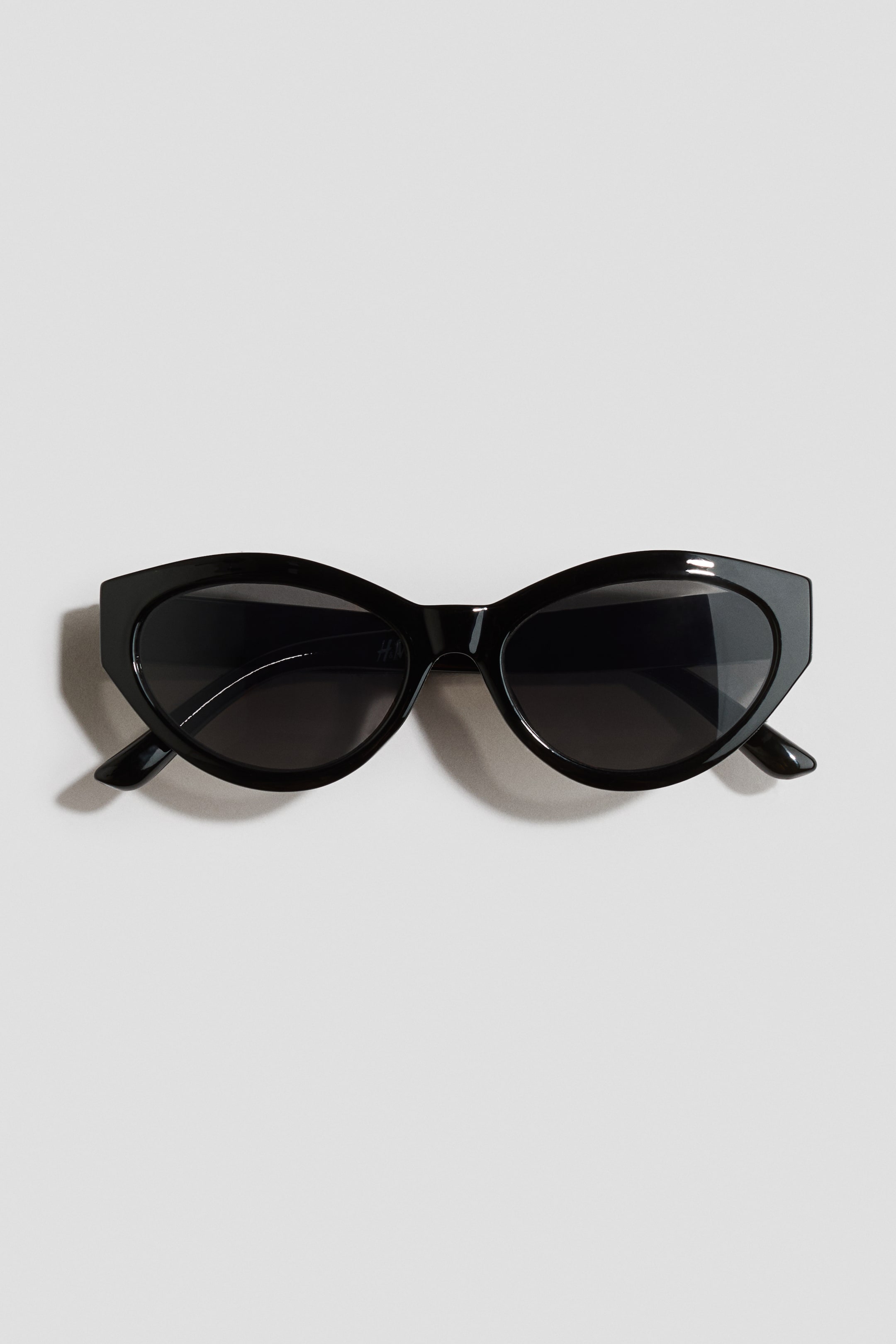 View larger image: Black cat-eye sunglasses showcase thick, glossy frames paired with dark, oval lenses. The frames boast subtly pointed outer corners, complemented by slender temples extending gracefully from the sides.