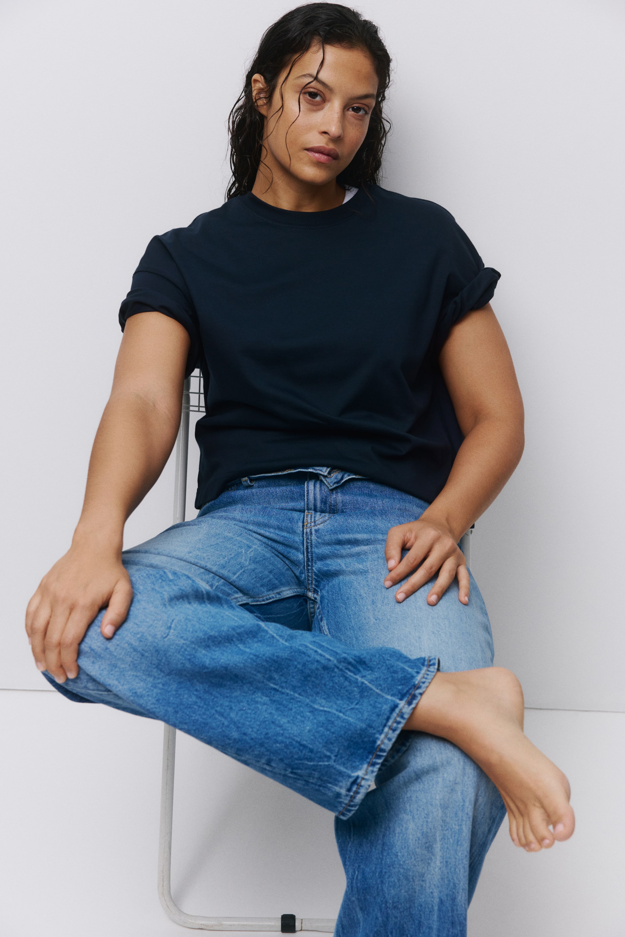 Vis større bilde: En mørkeblå t-skjorte med rund hals har oppbrettede korte ermer og en avslappet passform. Den er kombinert med mellomblå jeans som har en avslappet, rett benform med lett falming.