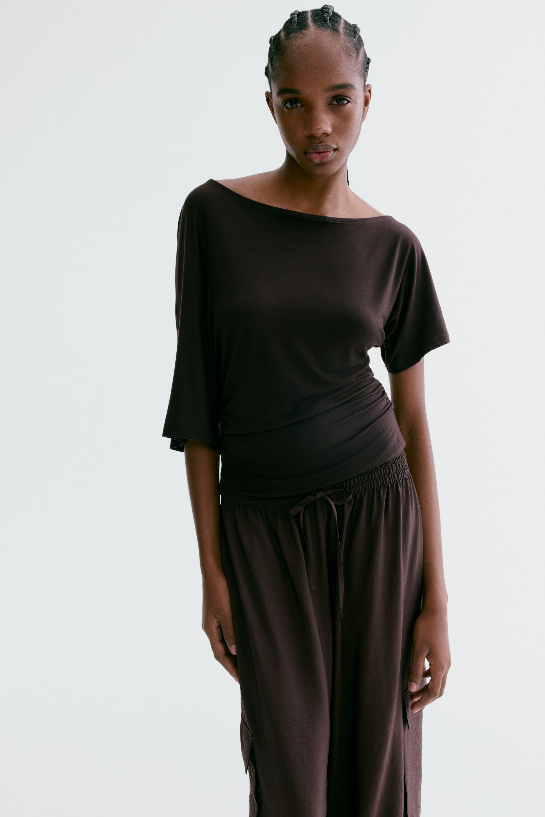 Visa större bild: Det mörkbruna setet har en båthalsad topp med korta, lösa dolmanärmar och sidodrapering, kombinerat med löst sittande byxor som har en elastisk dragsko i midjan och en sidoficka. Outfiten är något vänd åt höger.