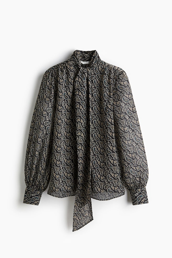 Un chemisier en chiffon transparent, vu de face, présente un fond sombre avec un motif cachemire répétitif en or et vert atténués. Il est conçu avec un col à nœud et une ouverture en forme de V à l'avant, avec des fronces se formant sur les épaules et de longues manches ballon se terminant par des poignets boutonnés. - 1