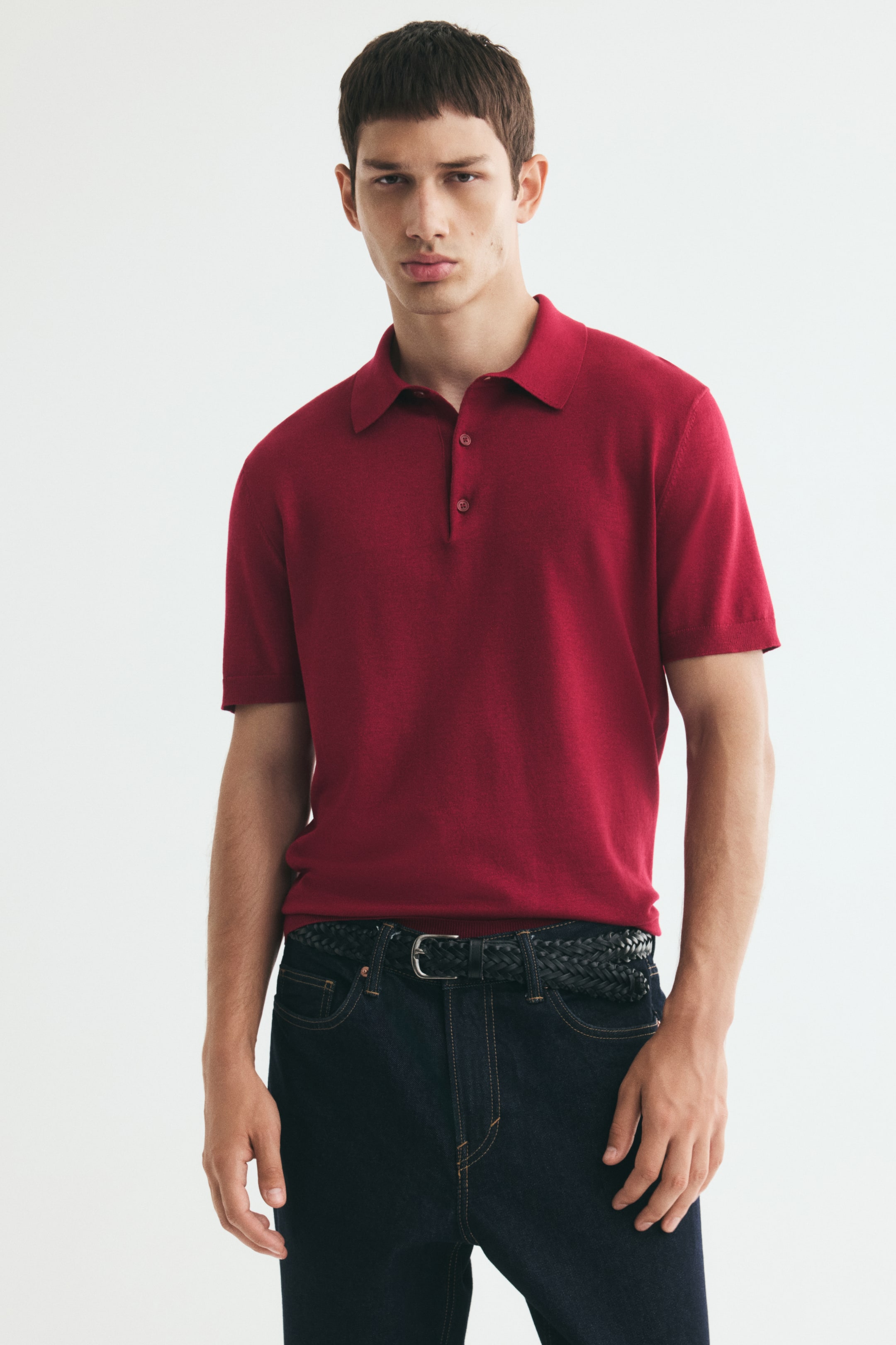 View larger image: The model is wearing a red knit polo shirt with a classic collar, button placket, and short sleeves. The shirt has a regular fit and is tucked into dark blue jeans, styled with a black braided belt.