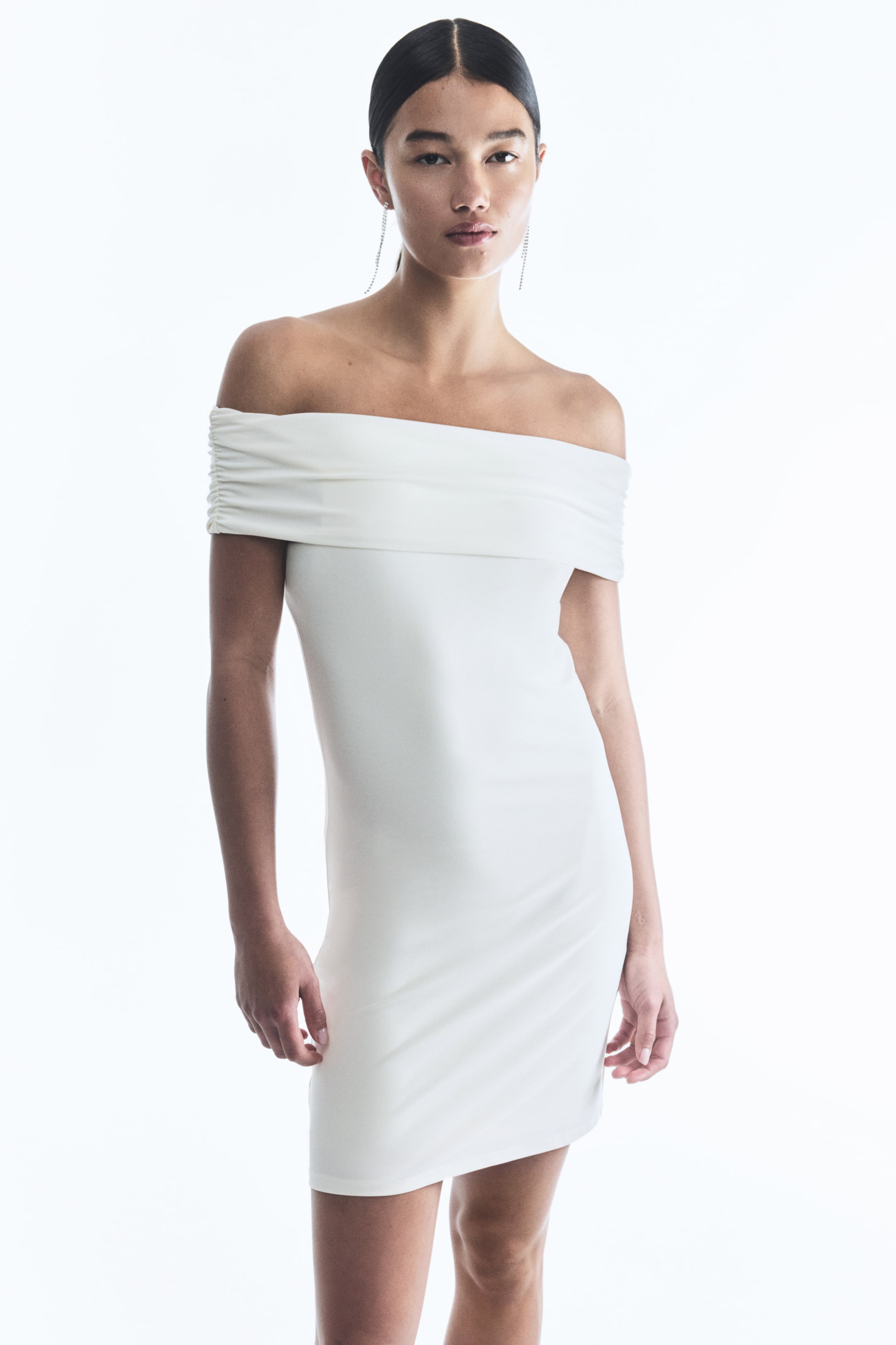 View larger image: A white off-the-shoulder mini dress is being presented from a front-facing angle, featuring a wide, gathered band across the bust. The dress is showcasing a fitted silhouette that is extending to mid-thigh.