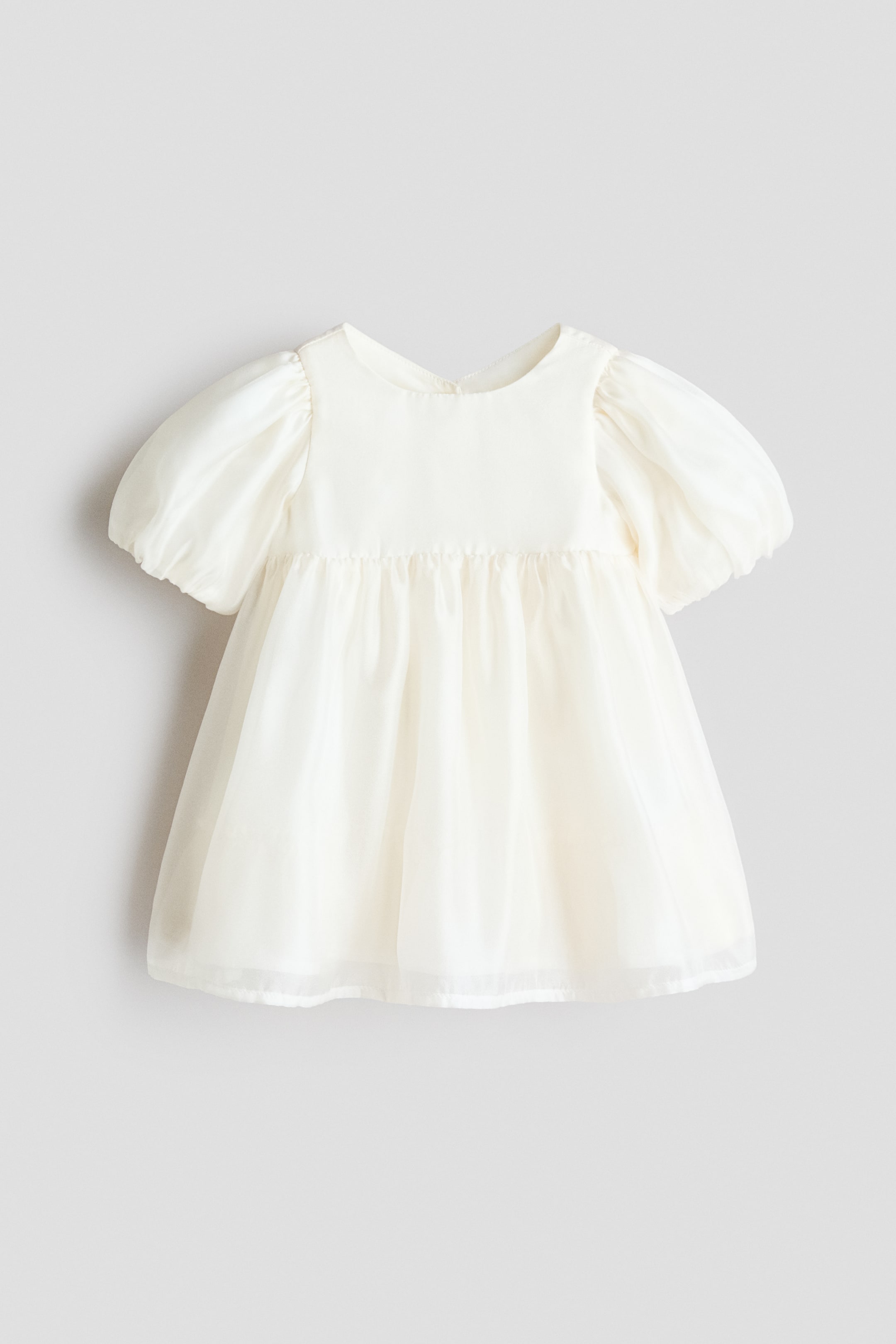 拡大画像を表示: オフホワイトのベビードレスは、ラウンドネックと短いパフスリーブを特徴としています。不透明なボディスは、ギャザーの入ったシアースカートに変化し、不透明な裏地の上に重ねられています。