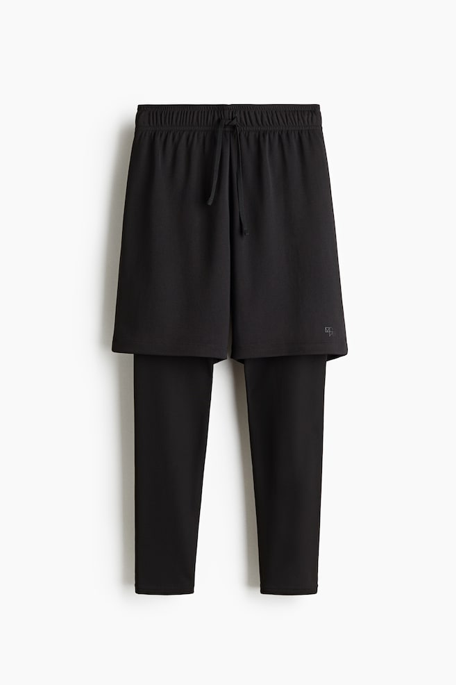 A black athletic garment, presented from the front, is featuring an integrated design of shorts layered over full-length leggings. The shorts are showing an elasticized waist with a visible drawstring and a subtle logo on the lower right, while the leggings are extending to the ankles. - 1