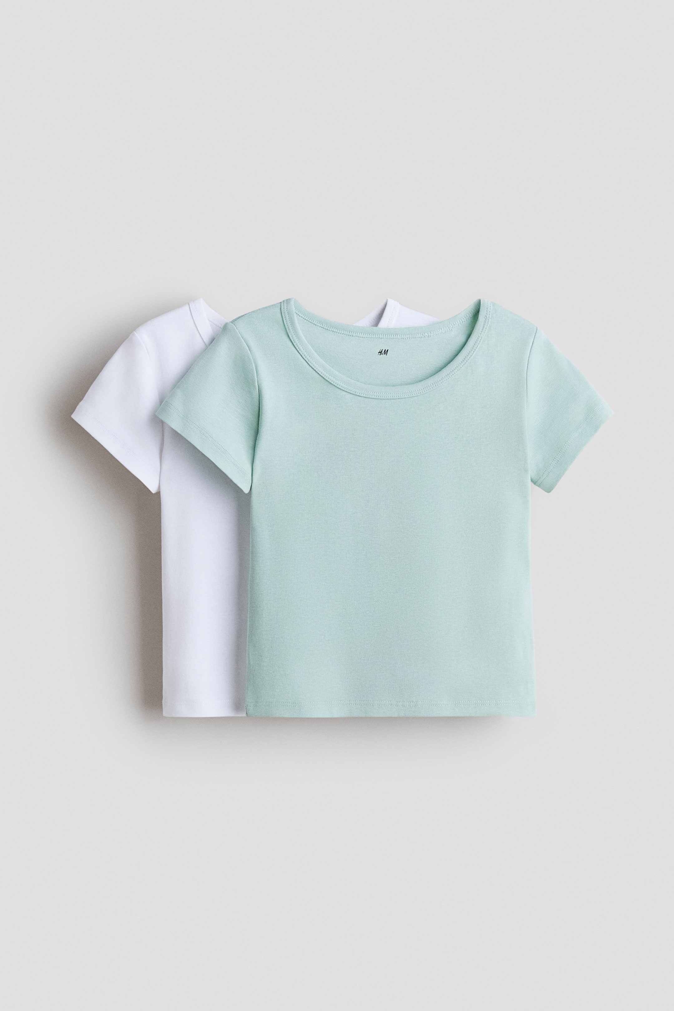 Zobrazit větší obrázek: Dva trička s krátkým rukávem jsou prezentována čelem dopředu, přičemž světle mátově zelené tričko je navrstveno přes bílé. Obě trička, vyrobená z měkkého bavlněného úpletu, mají kulatý výstřih s žebrovaným lemem.