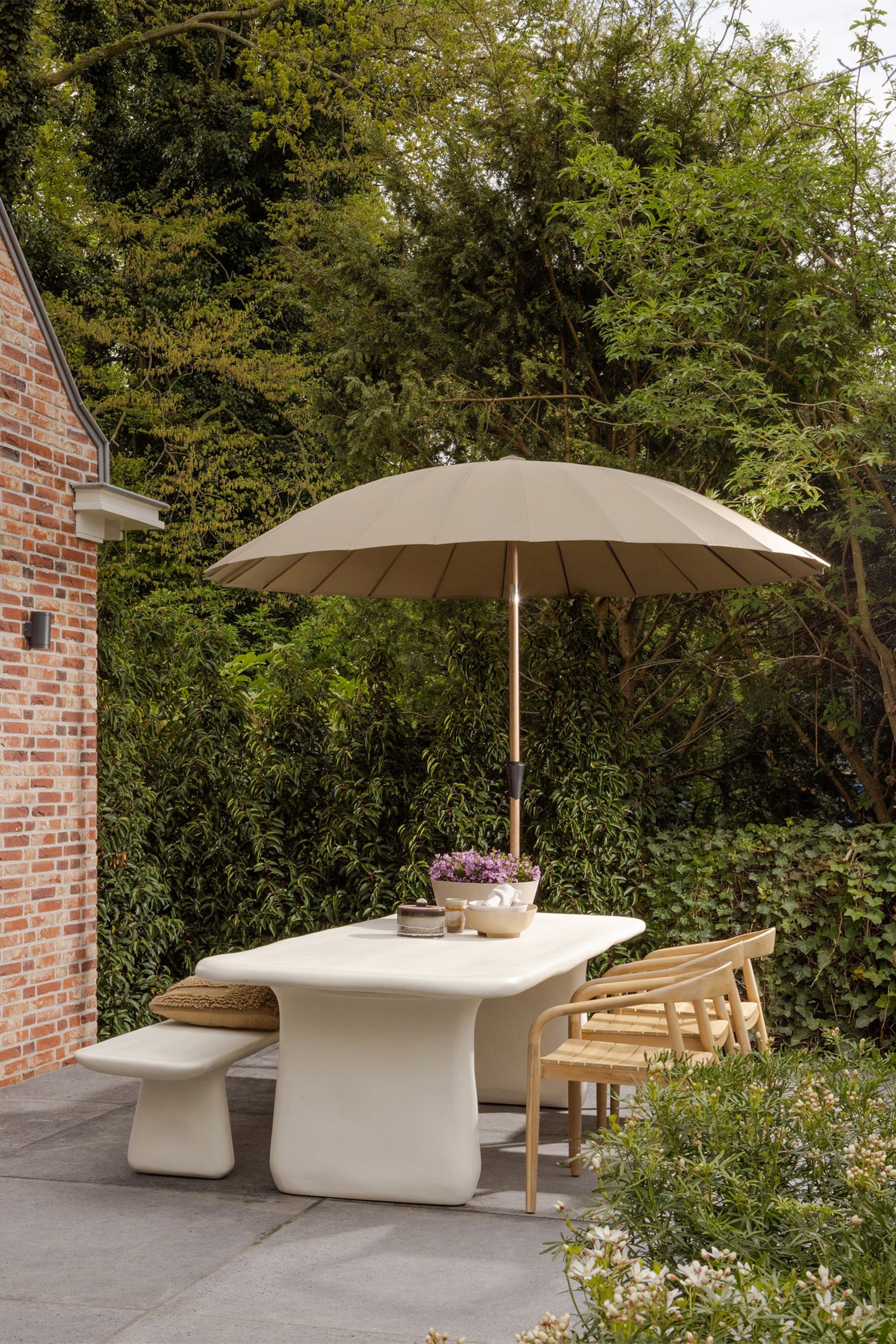 Agrandir l'image: Un ensemble de salle à manger extérieur présente une table rectangulaire de couleur crème avec des bords arrondis et deux grands pieds de socle courbés, accompagnée d'un banc solide assorti surmonté d'un coussin brun texturé. Deux chaises en bois clair avec des sièges à lattes et des dossiers courbés complètent l'ensemble, qui est ombragé par un grand parasol rond beige et vu sous un angle avant-droit.