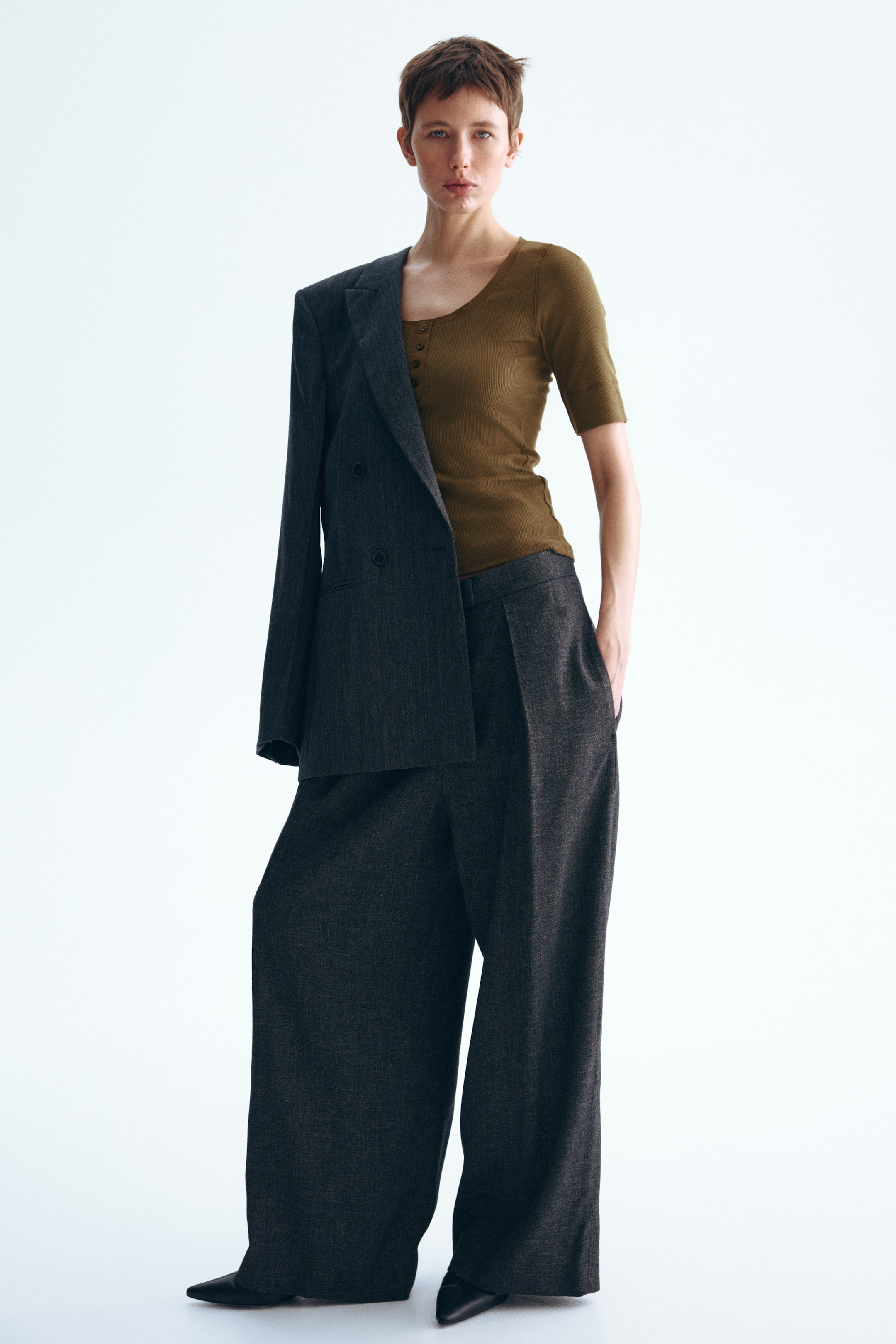 Grotere afbeelding bekijken: Een donkergrijze double-breasted blazer is nonchalant over één schouder gedrapeerd, waardoor een olijfgroene henley met korte mouwen zichtbaar wordt. Donkergrijze geplooide wijde broek en zwarte schoenen met puntige neus maken de outfit compleet.