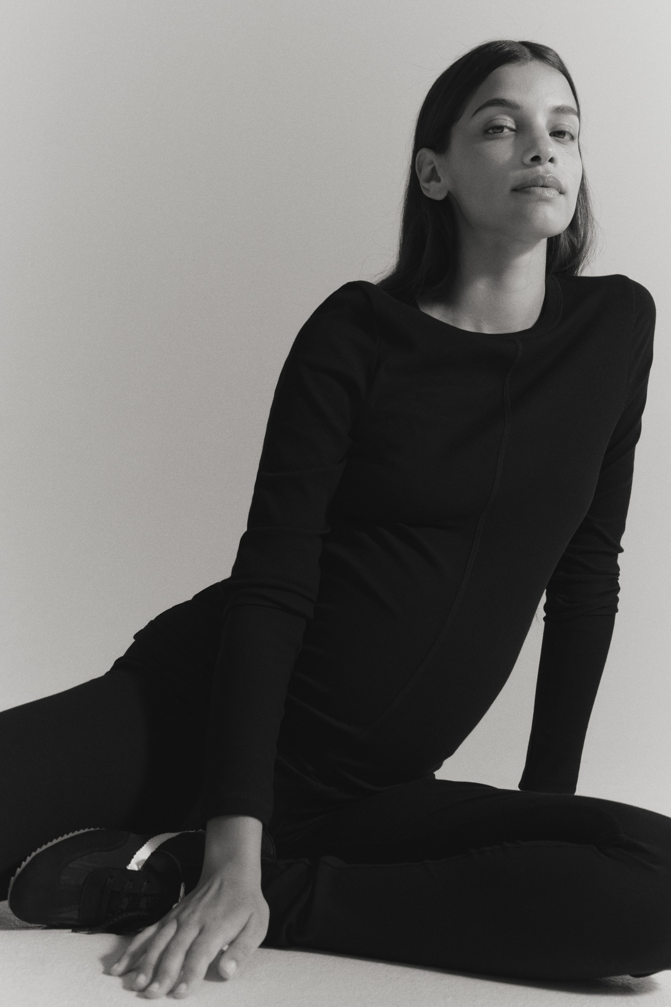 Se større billede: Modellen sidder på gulvet iført et sort sæt bestående af en ribstrikket jerseytop og leggings. Den tætsiddende top har rund halsudskæring og lange ærmer med synlige flatlock-sømme, kombineret med matchende ribstrikkede leggings.