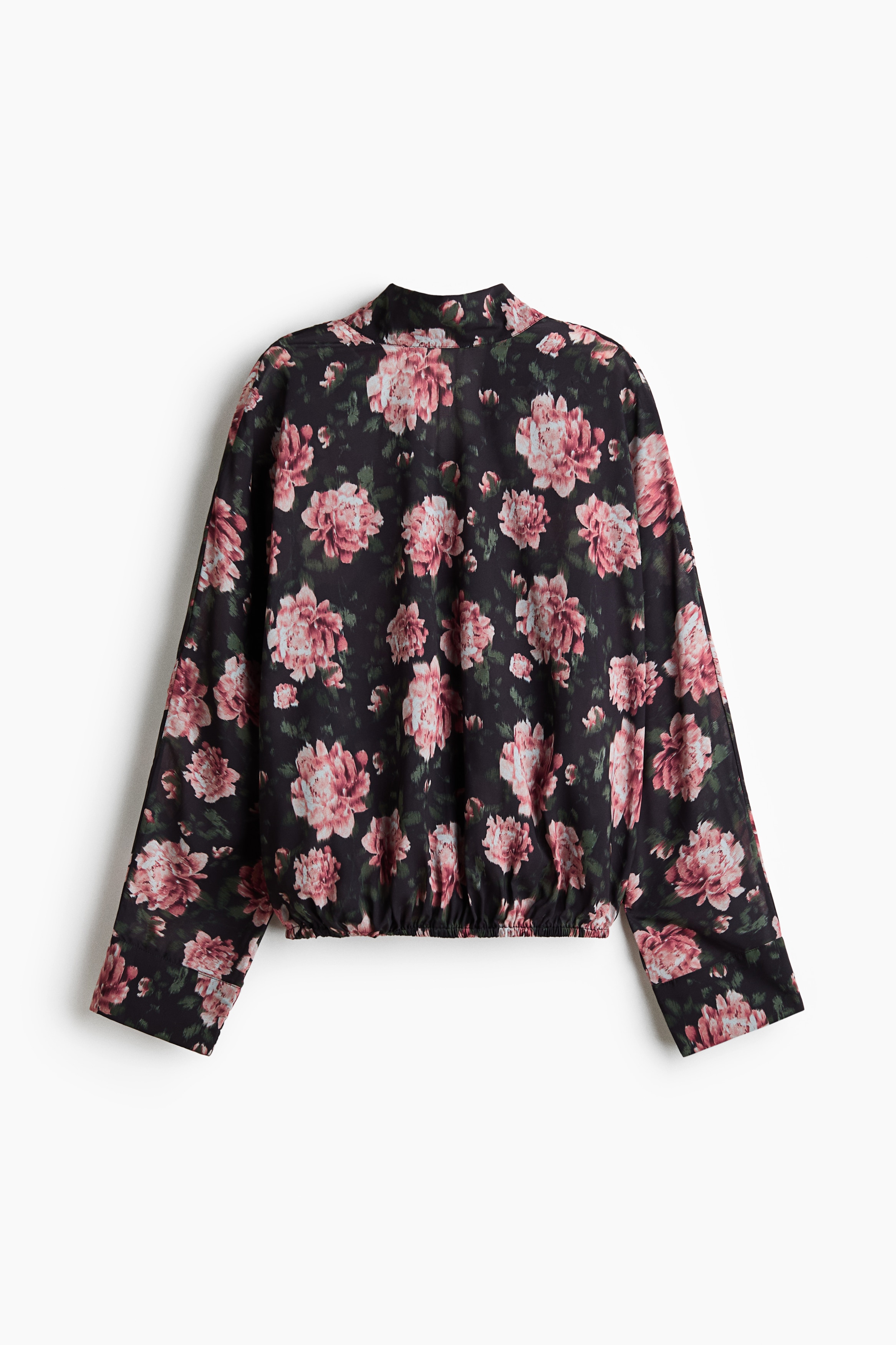 De model draagt een blouse met bloemenprint in een donkere, transparante weving, met een hoge bandkraag en linten die langs de achterkant naar beneden hangen. De blouse heeft verlaagde schouders, lange mouwen met manchetten en een elastische zoom, gestyled met olijfgroene trekkoordbroeken.