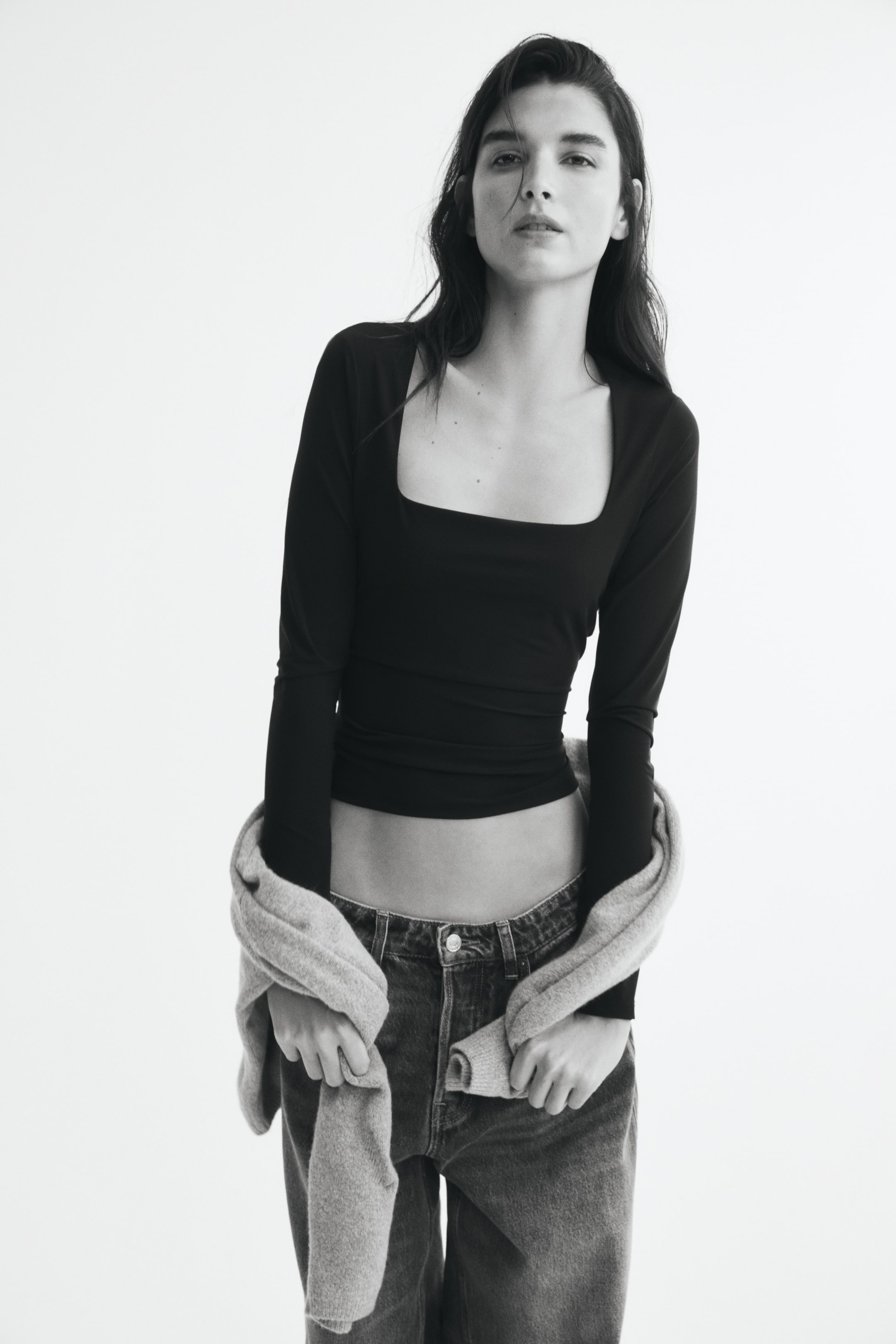 Büyük resmi görüntüle: Modelin üzerinde derin, kare yaka ve uzun kollara sahip, vücuda oturan siyah bir üst bulunuyor. Üst, koyu renk denim jean ile kombinlenmiş; model, kollarını tutarak beline açık renkli bir kazak bağlamış.