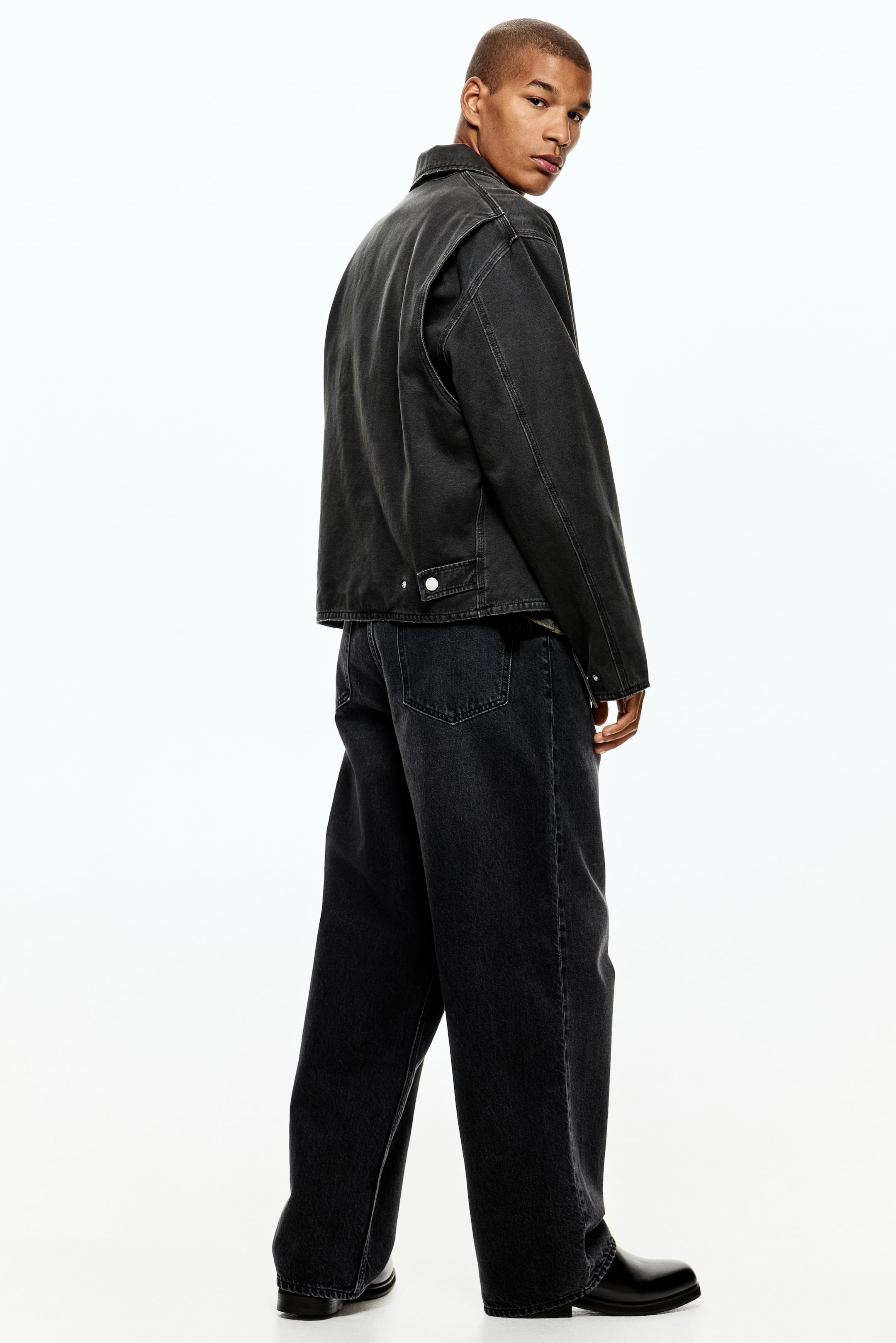 Ampliar la imagen: Una chaqueta de trabajo gris oscuro está siendo presentada desde la parte trasera y el lado derecho, con un destacado contraste de costuras blancas, un cuello en punta y dos botones de metal blanco en el dobladillo trasero. Se está combinando con jeans de pierna ancha gris oscuro a juego, que muestran un ajuste suelto y bolsillos traseros tipo parche.