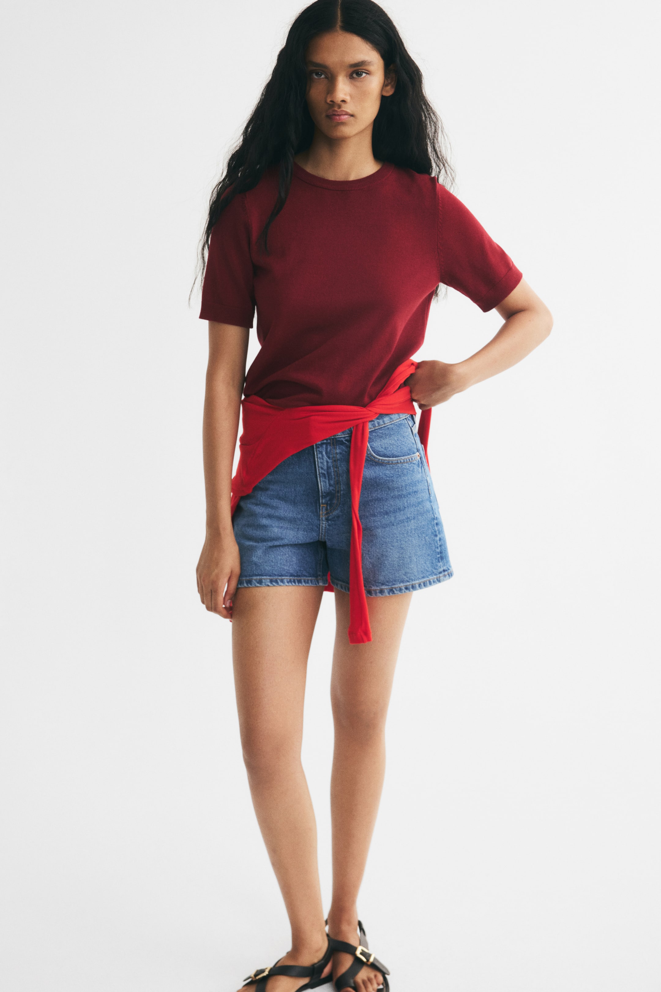 View larger image: The model is wearing a dark red knitted short-sleeved crew-neck top, paired with blue denim shorts and black flat sandals. A red long-sleeved top is tied around the model’s waist, and the model is holding the fabric.