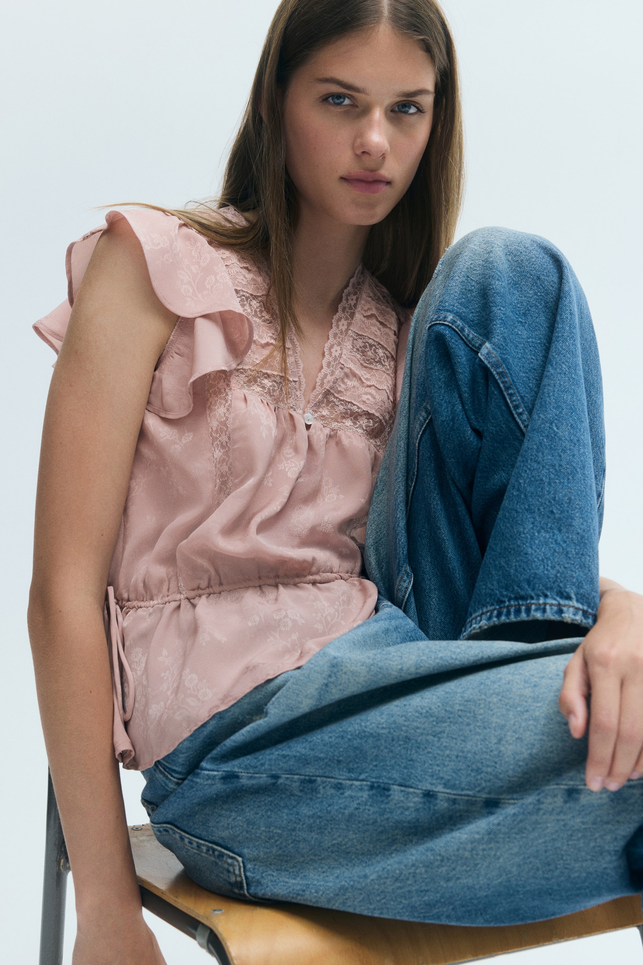 View larger image: The model is wearing a pink, sheer-weave blouse featuring a lace-trimmed V-neckline, flutter sleeves, and a gathered drawstring waist, paired with blue denim jeans.