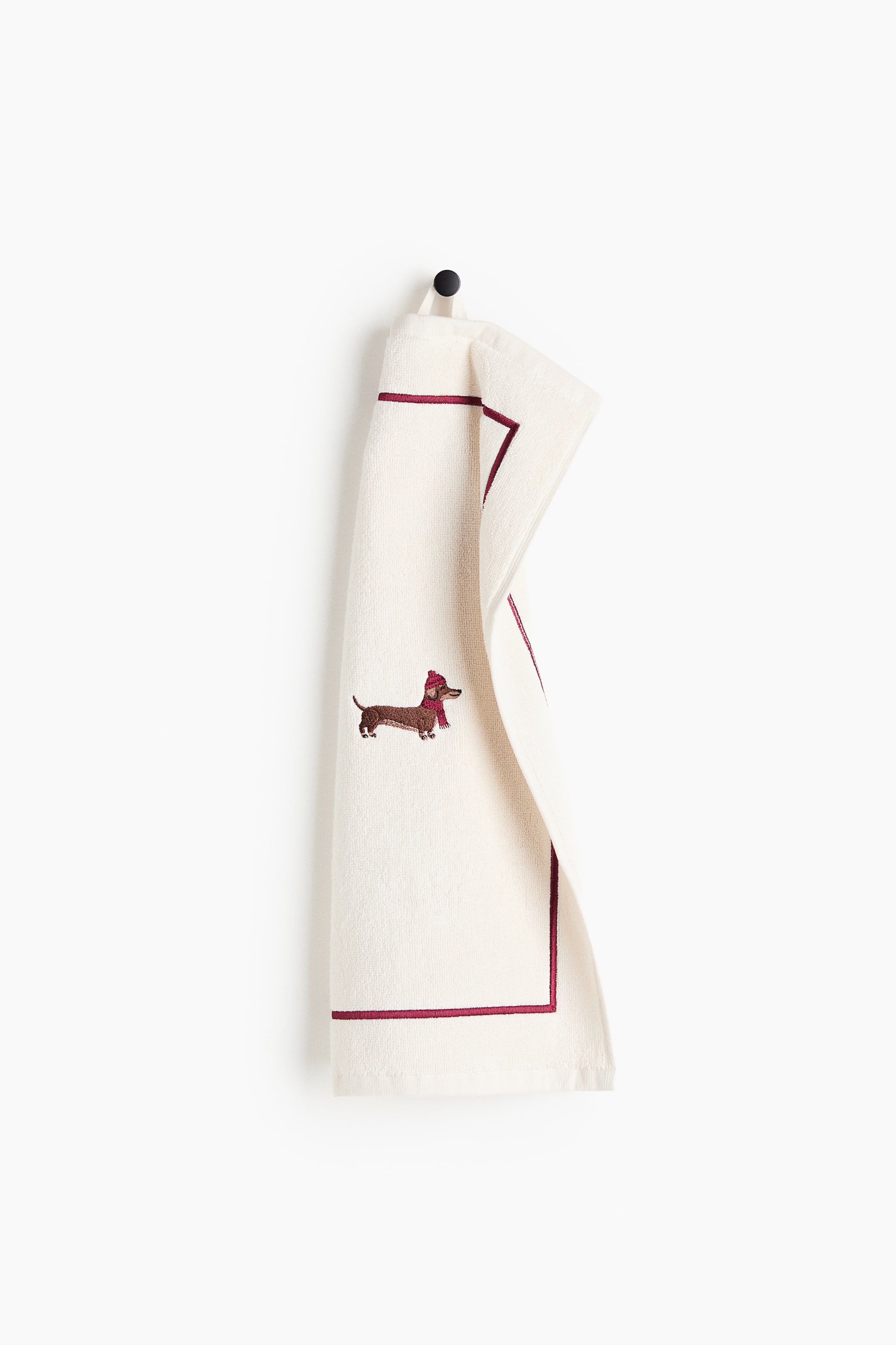 View larger image: A cream-coloured terry cloth hand towel is hanging from a top loop, displaying its front design with a thin burgundy embroidered border. An embroidered dachshund wearing a red knit hat and scarf is featured near the centre of the towel.
