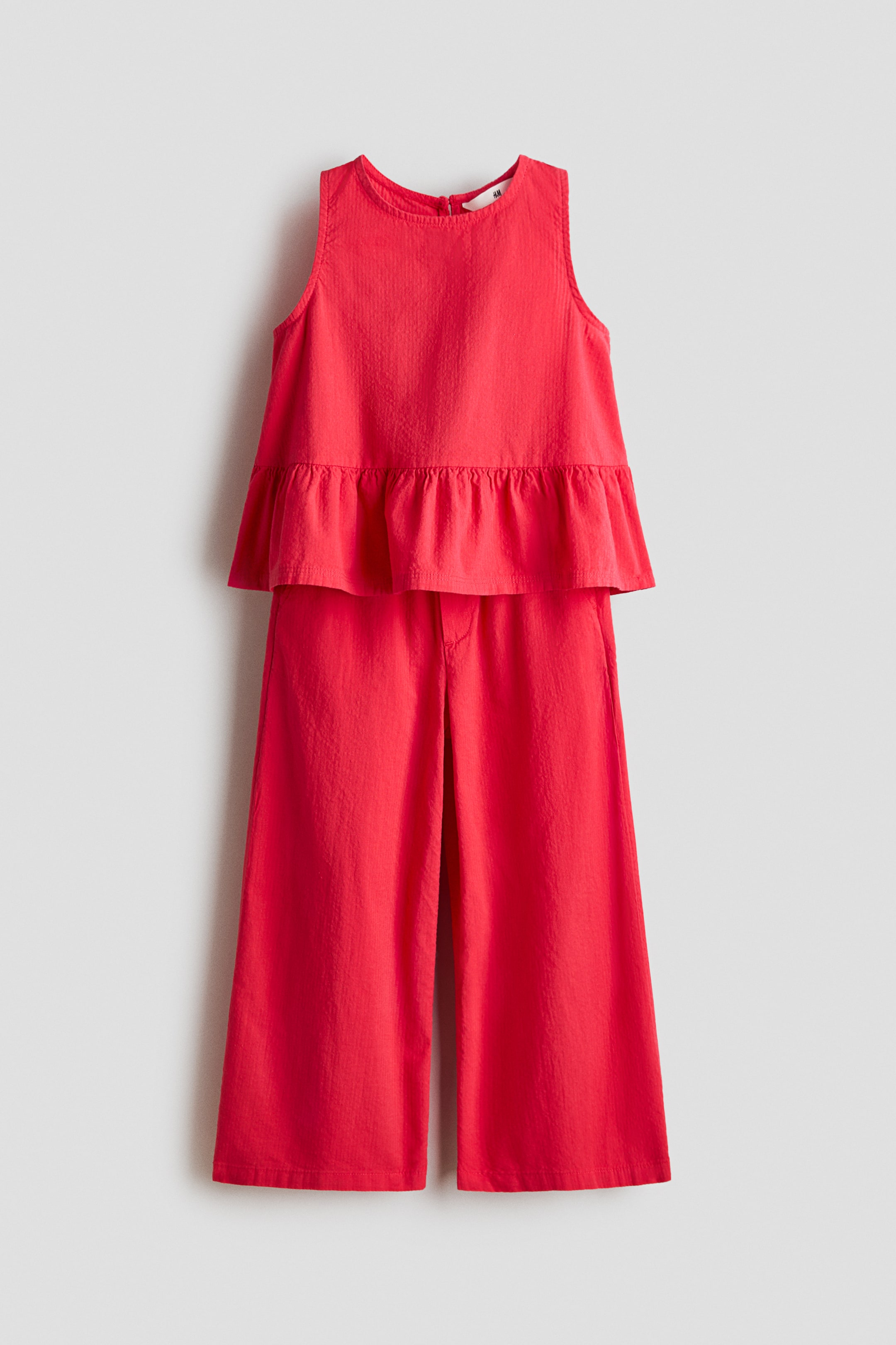 View larger image: A red two-piece outfit is shown from the front, featuring a sleeveless top and wide-leg trousers. The top has a round neckline and a gathered peplum hem, while the cropped trousers offer a relaxed fit. Both pieces are crafted from a subtly textured fabric.