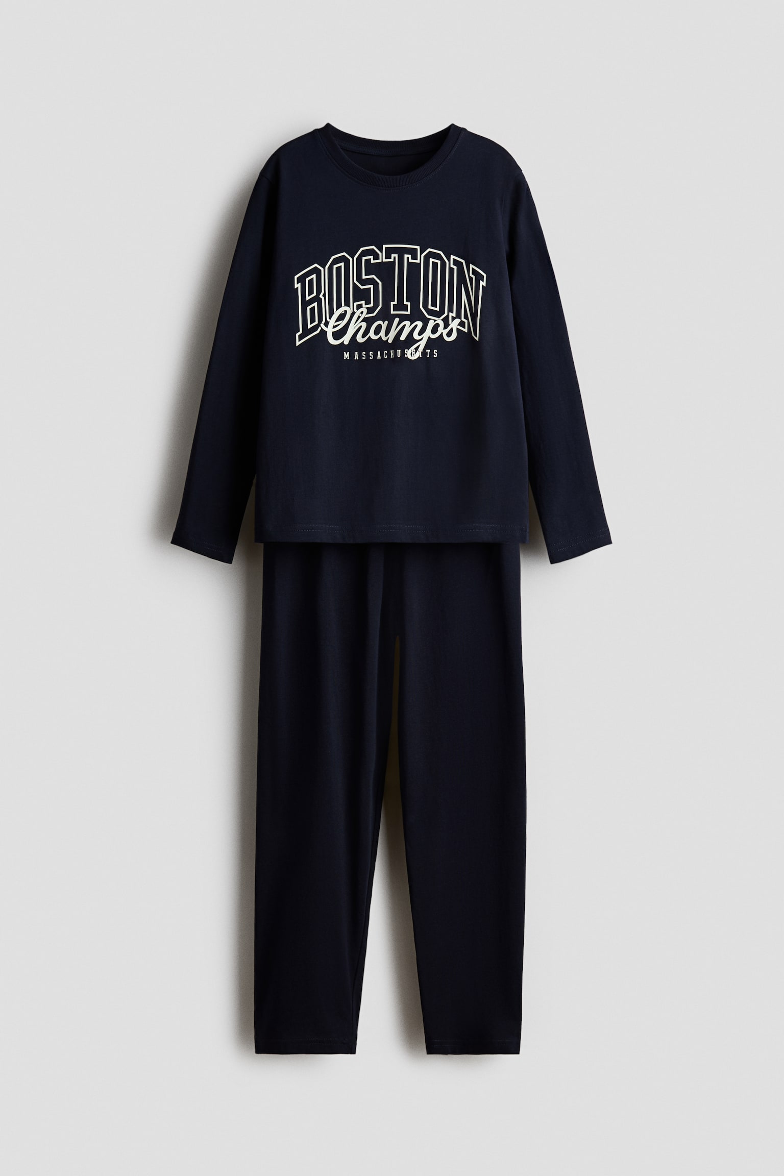 Se muestra de frente un conjunto de pijama azul marino oscuro, compuesto por una parte superior de manga larga con cuello redondo y un pantalÃ³n recto a juego. La parte superior presenta un estampado grÃ¡fico blanco en el pecho con el texto "BOSTON Champs MASSACHUSETTS" en estilo universitario.