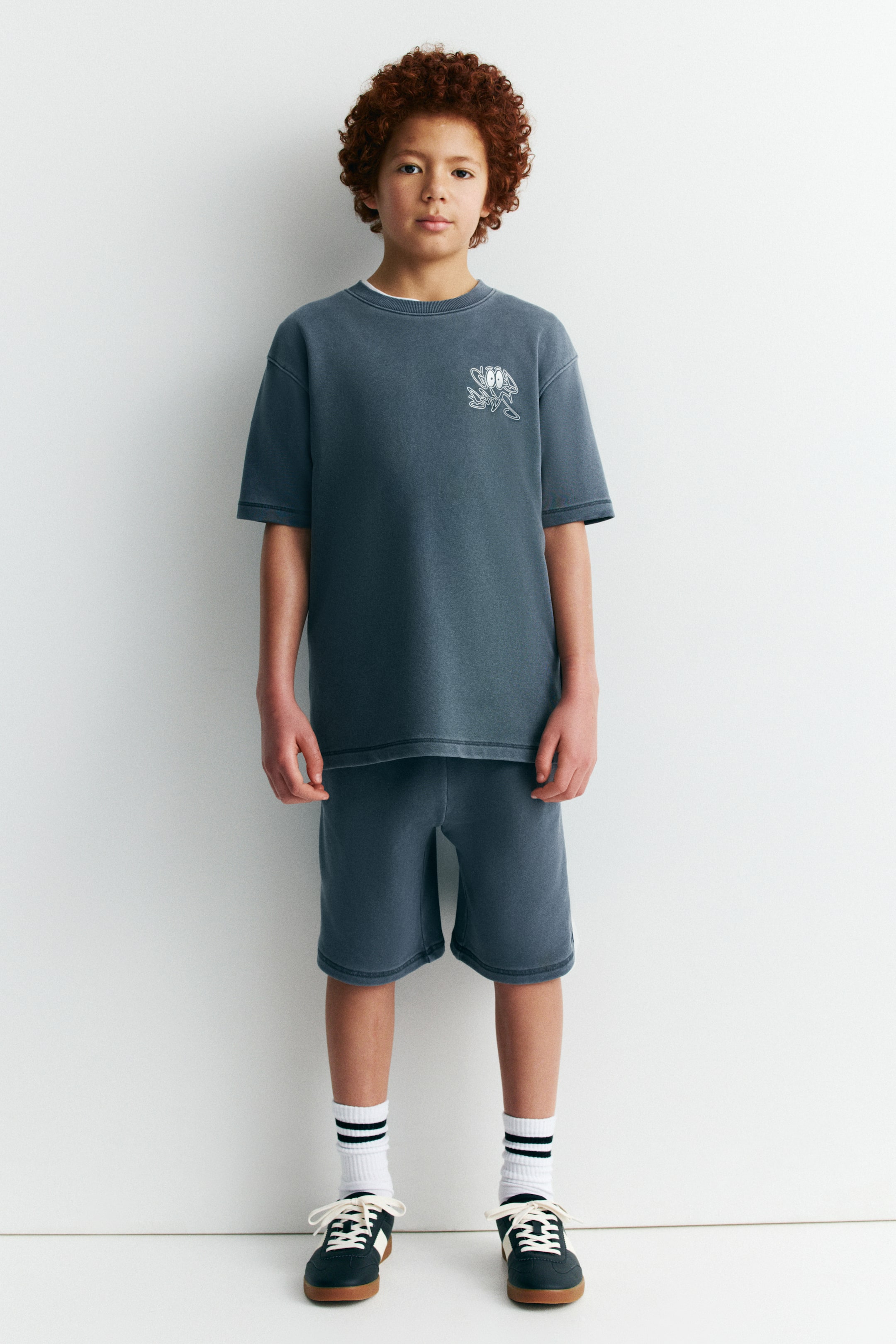 Ver imagen más grande: Un conjunto de camiseta de manga corta y pantalones cortos a juego en color gris carbón lavado se presenta de frente. La camiseta cuenta con hombros caídos y un pequeño estampado gráfico blanco en el pecho izquierdo, mientras que los pantalones cortos de ajuste relajado llegan por encima de la rodilla con costuras visibles en el dobladillo.