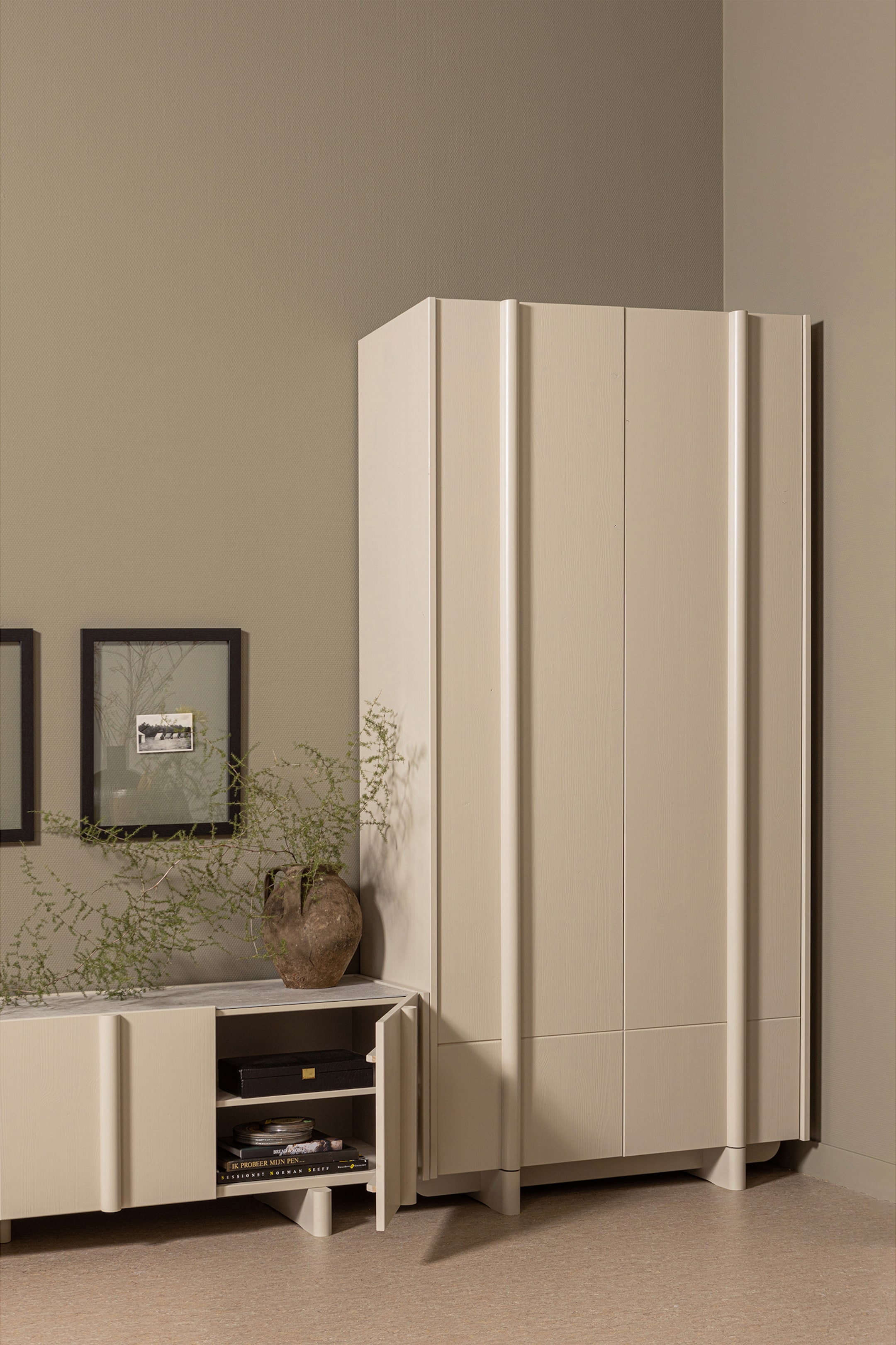 Größeres Bild ansehen: Ein blasses beige modulares Schranksystem präsentiert seine Vorder- und rechte Seite. Das System besteht aus einem hohen viertürigen Schrank mit vertikalen geriffelten Paneelen an den äußeren Kanten, der neben einem kürzeren dreitürigen Schrank steht, dessen eine Tür geöffnet ist und die Inneneinrichtung mit Regalen zeigt. Beide Schränke ruhen auf zurückgesetzten Sockeln.