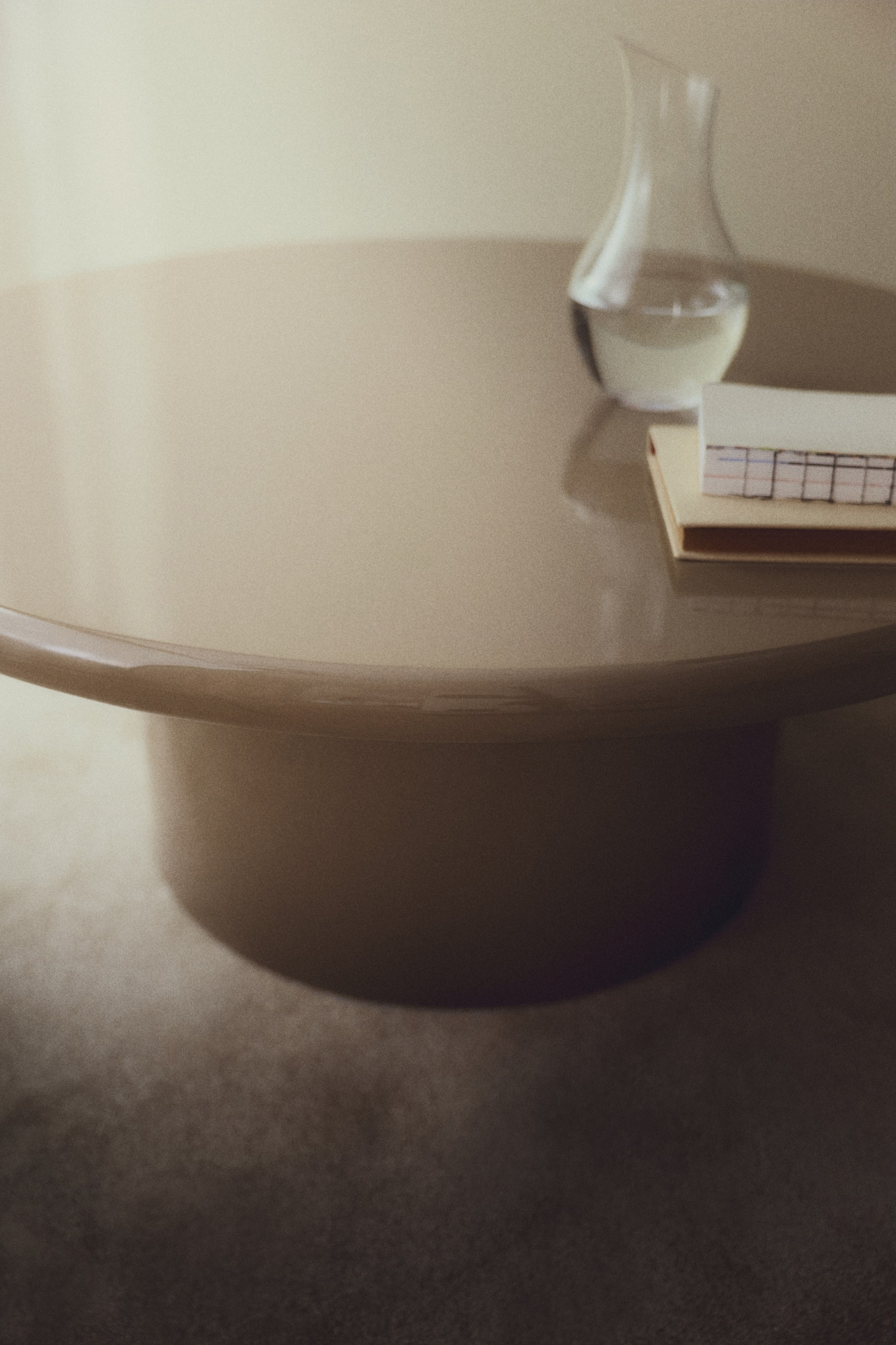 Agrandir l'image: Une table basse ronde présente une surface lisse et réfléchissante de couleur taupe avec un bord légèrement surélevé. Elle est soutenue par une base cylindrique épaisse, et une carafe en verre transparent ainsi qu'une pile de petits carnets reposent sur sa surface.