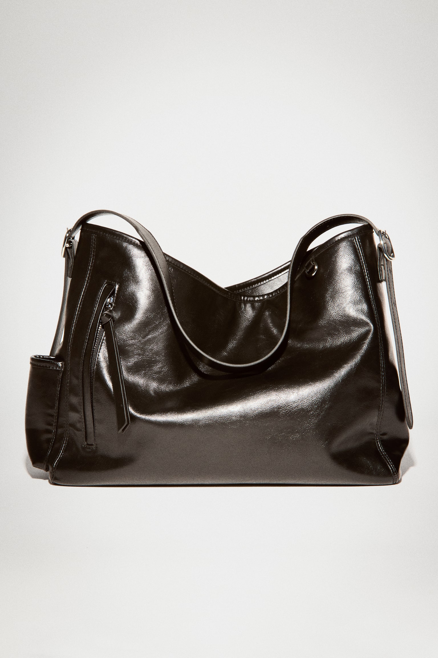 A black leather shoulder bag is standing upright and facing forward, showing a glossy, textured finish. A single flat shoulder strap arches over the curved top, and a vertical zippered pocket with a long pull tab details the front left side. - 1