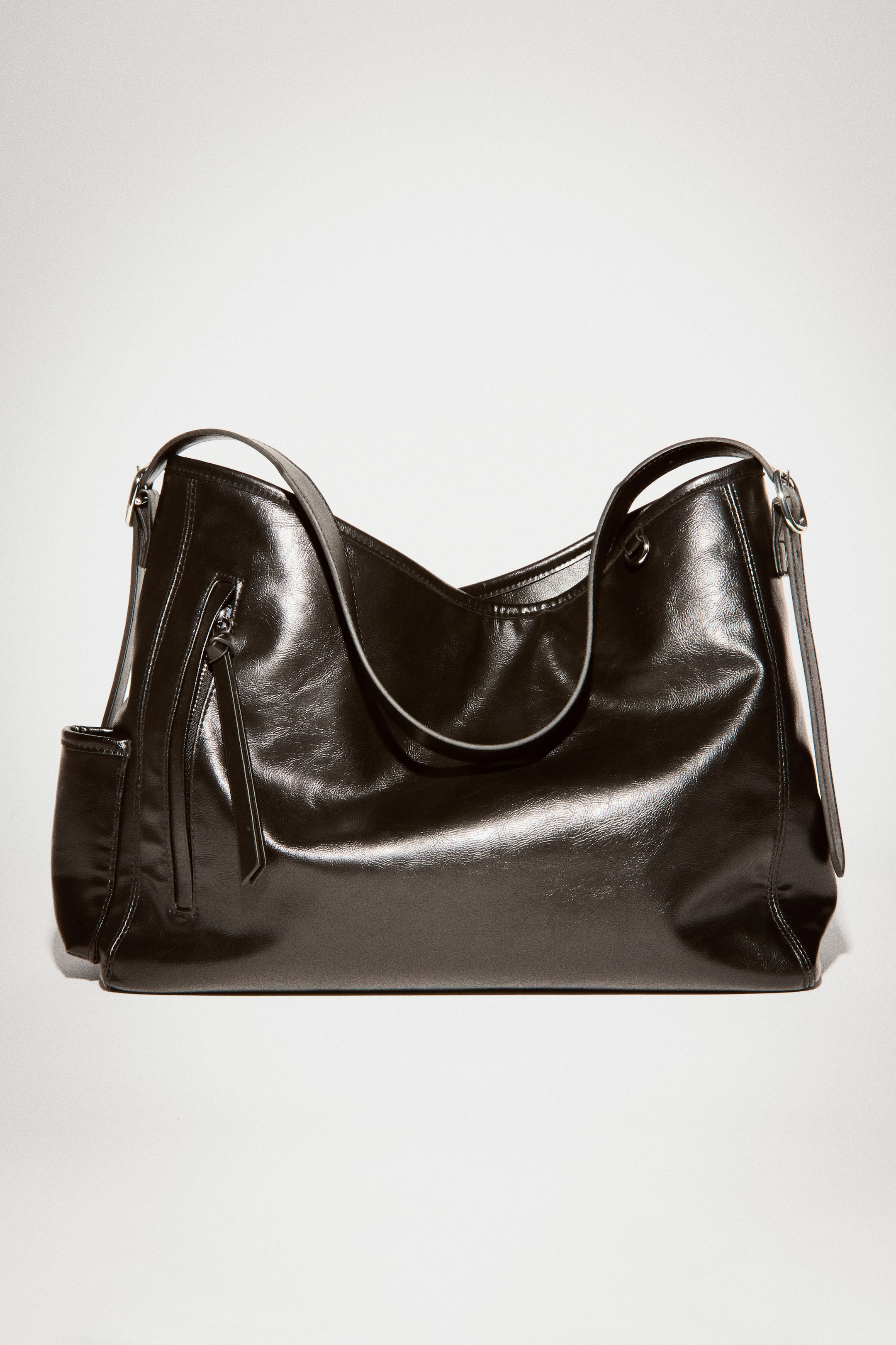 Visualizza l'immagine ingrandita: Una borsa a spalla nera in tessuto spalmato lucido è in posizione verticale, vista frontale, e mette in risalto una silhouette morbida e rilassata e una tracolla regolabile fissata con fibbie metalliche. La borsa presenta una tasca esterna verticale con zip sul davanti, con un lungo tirazip annodato, e una tasca aperta a fessura sul lato sinistro.
