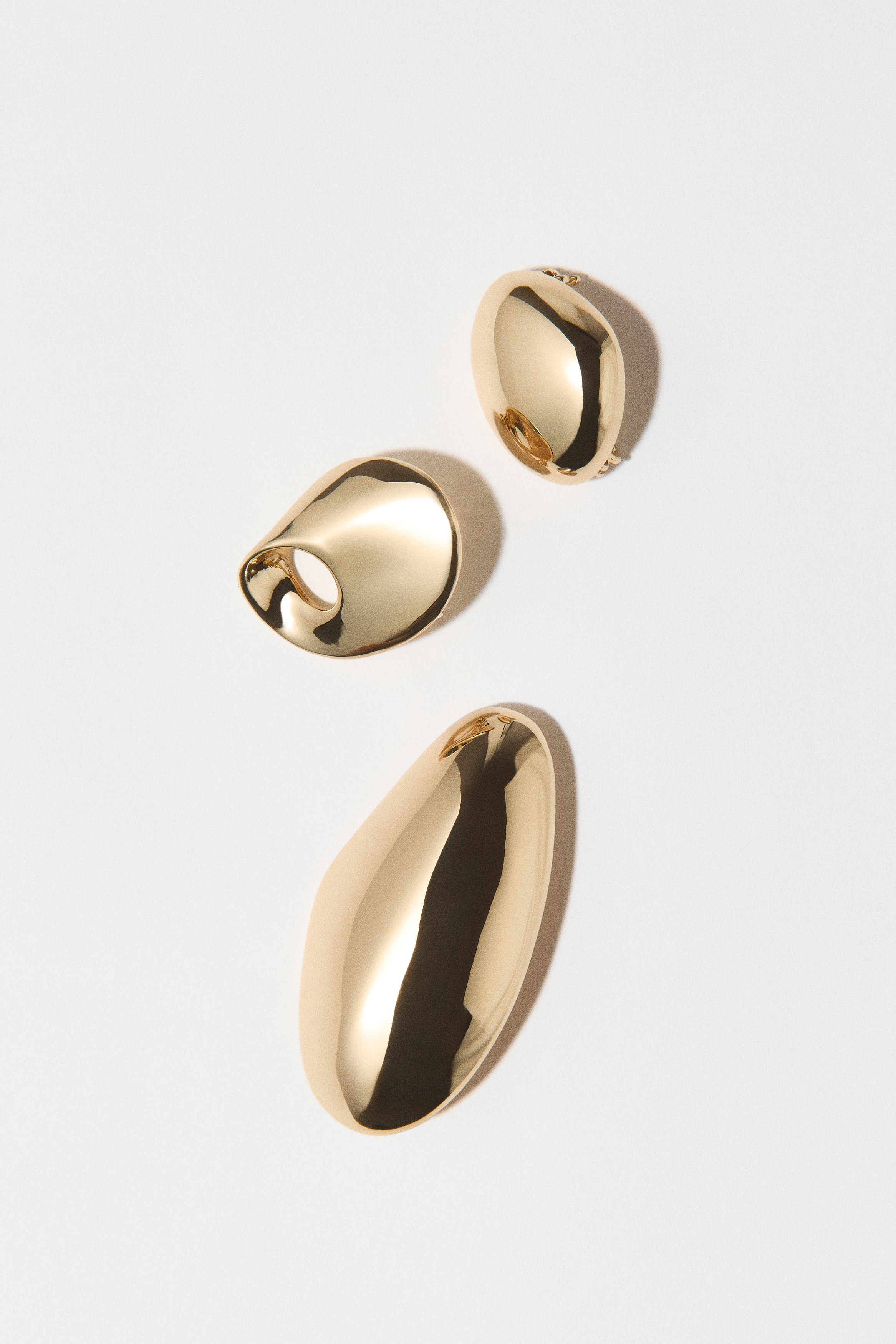 A set of three gold-toned, polished metal accessories showcases smooth, organic, pebble-like shapes with a highly reflective finish. The pieces are arranged vertically, featuring a small rounded form at the top, a middle piece with an off-centre circular cut-out, and a larger, elongated curved piece at the bottom.