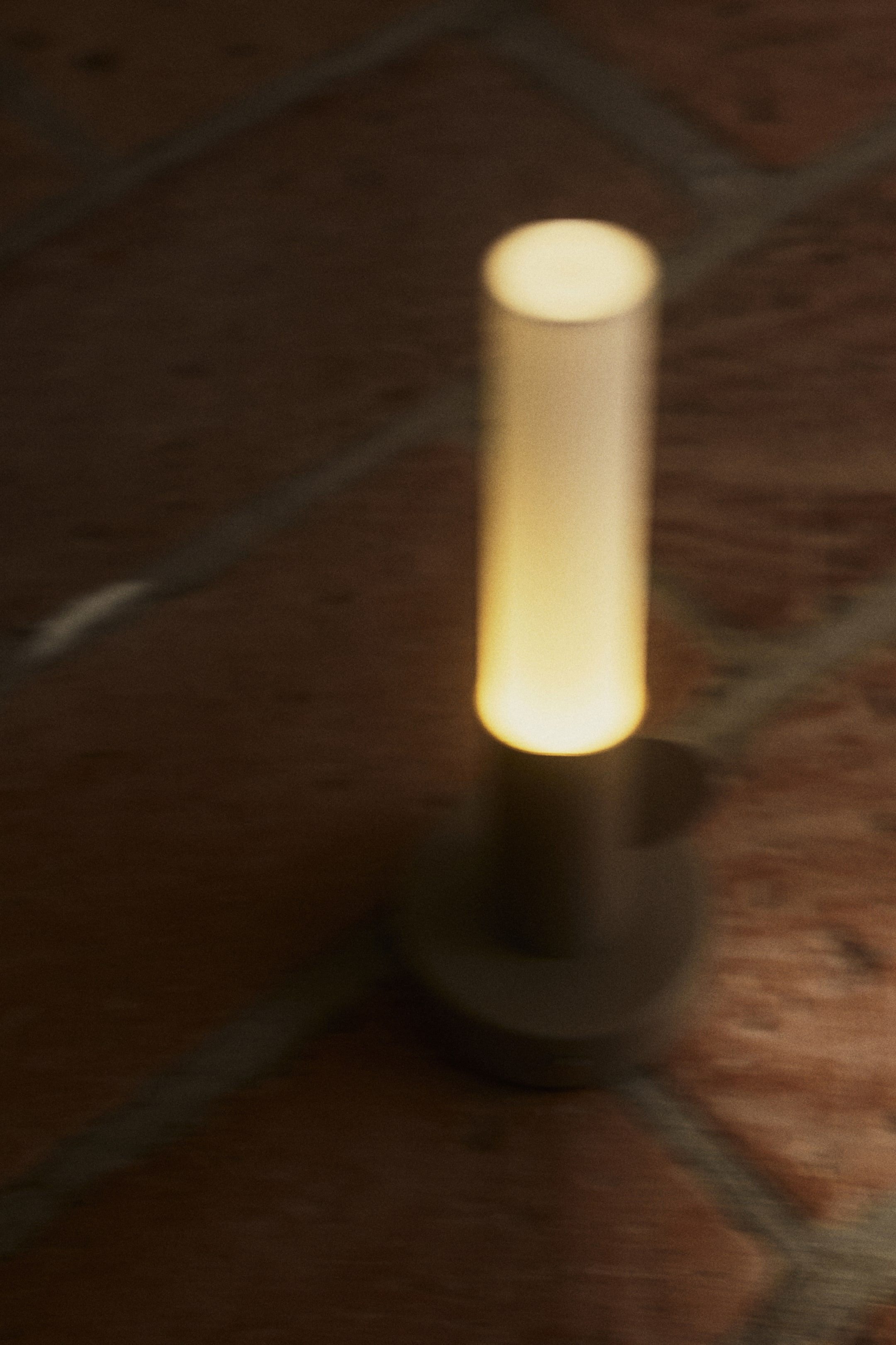 View larger image: A cylindrical light stands upright, glowing with a warm yellow illumination. It features a translucent top section and a dark, circular base.