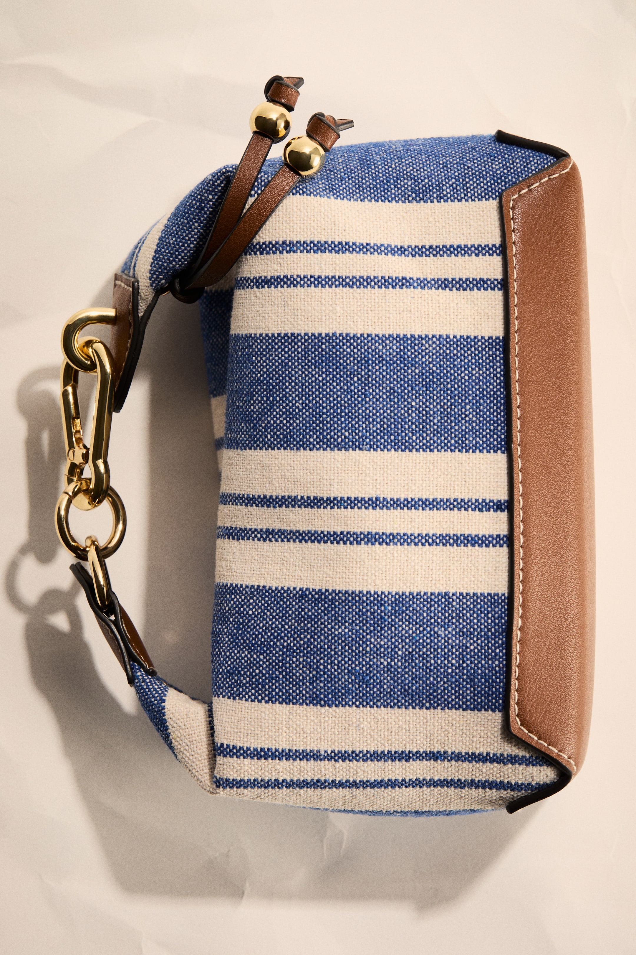 View larger image: The bag features blue and off-white horizontal stripes on a woven fabric body, accented with light brown leather trimming along the right side and bottom edge. Gold-tone metal hardware, including a carabiner-style clasp and two decorative beads on thin leather straps, adorns the top left section of the bag.