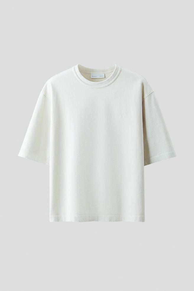 Ein cremefarbenes, kastiges T-Shirt wird von der VORDERSEITE präsentiert, mit einer lockeren Passform, überschnittenen Schultern und einem runden, gerippten Ausschnitt. Ein kleiner gestickter Buchstabe erscheint auf der Brust, und es ist mit einem geraden Saum abgeschlossen. - 1