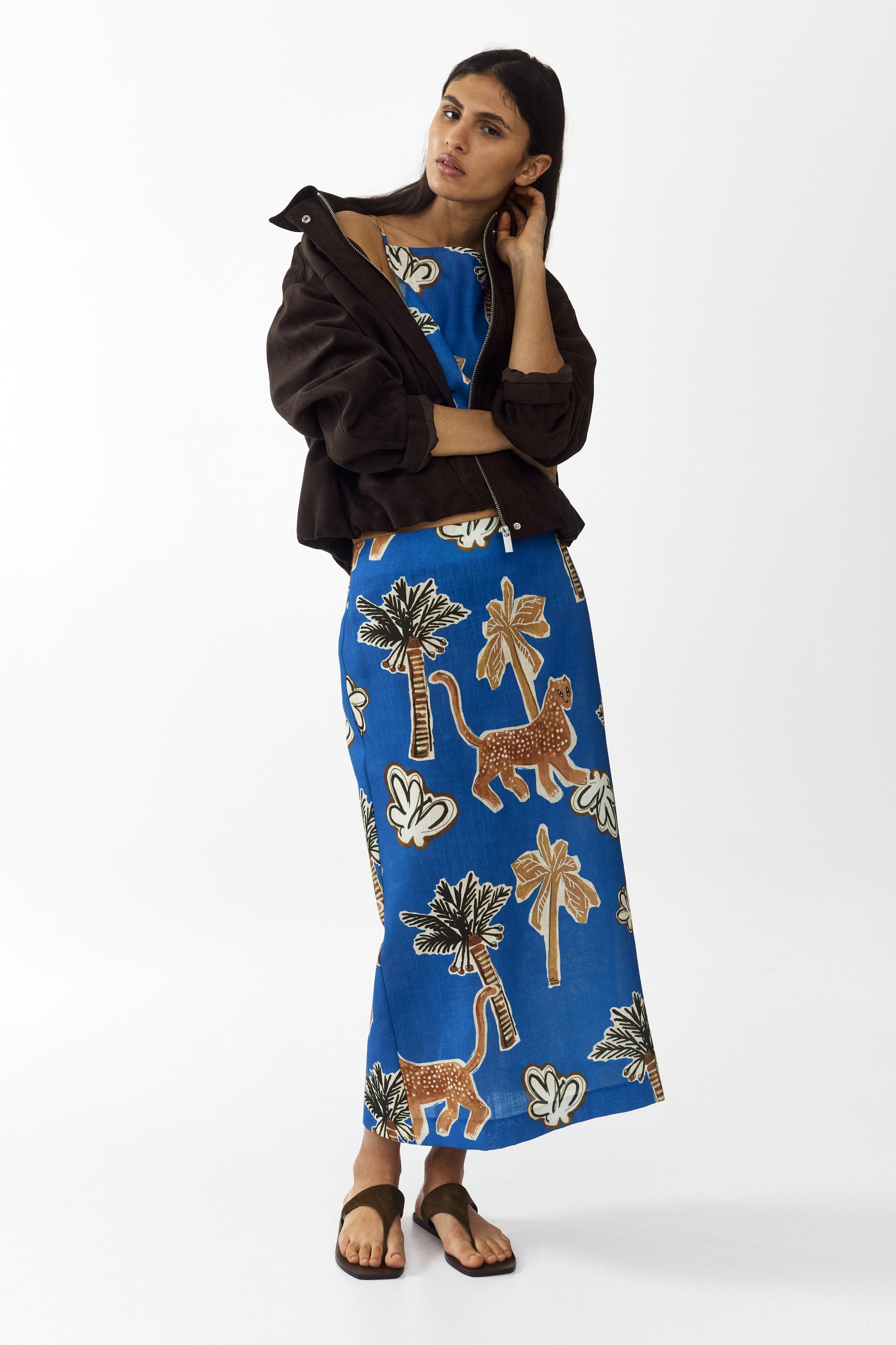 Visualizza immagine più grande: Una giacca corta marrone scuro è indossata aperta sopra un completo blu a due pezzi, dove un top con spalline e una lunga gonna mostrano entrambi un motivo di palme e animali maculati. Sandali infradito marrone scuro completano l'insieme.