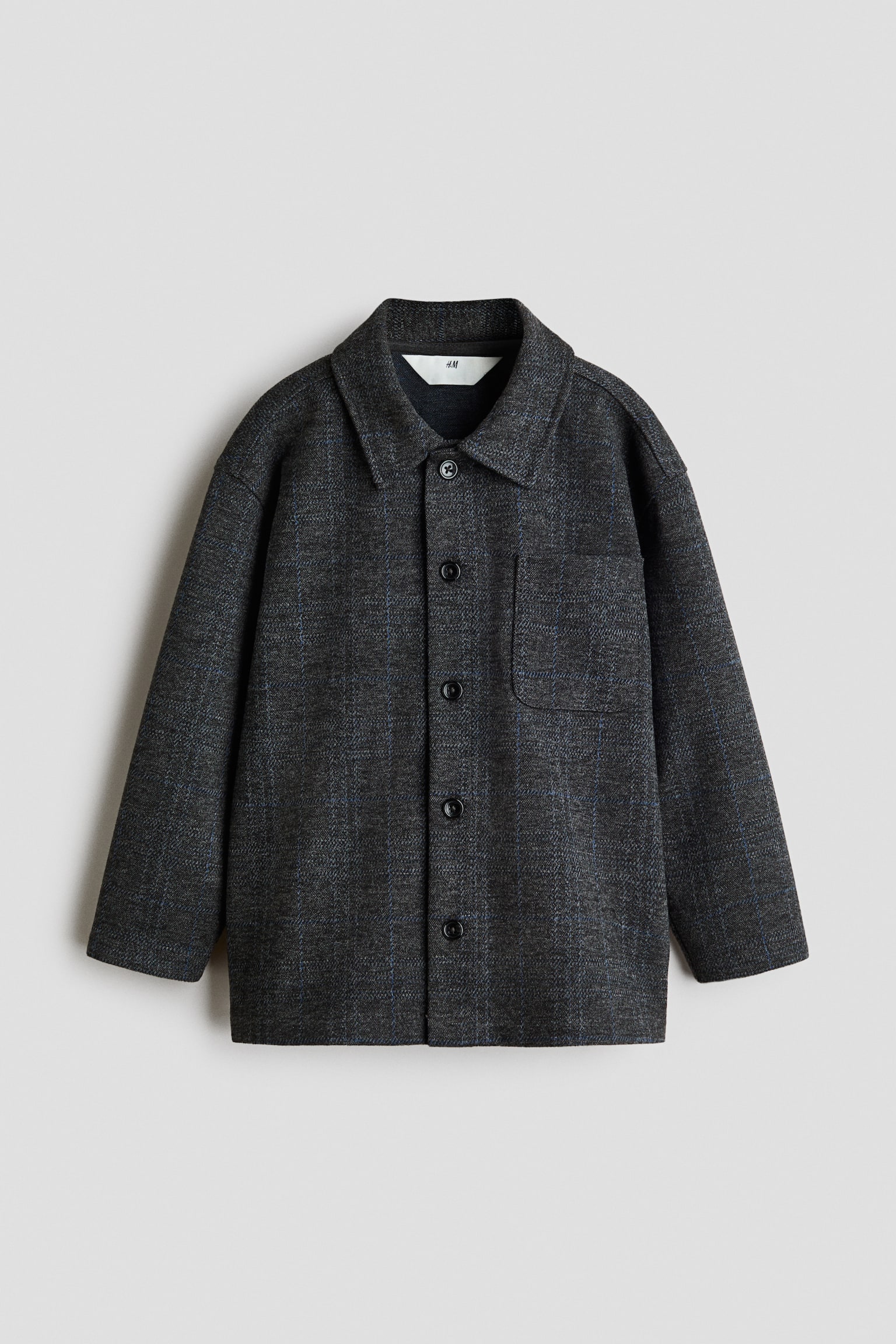 A loose-fit overshirt in dark grey textured jersey is shown from the front, featuring a subtle blue grid pattern. It features a classic collar, button-down front, a single chest patch pocket, and dropped shoulders with long sleeves.