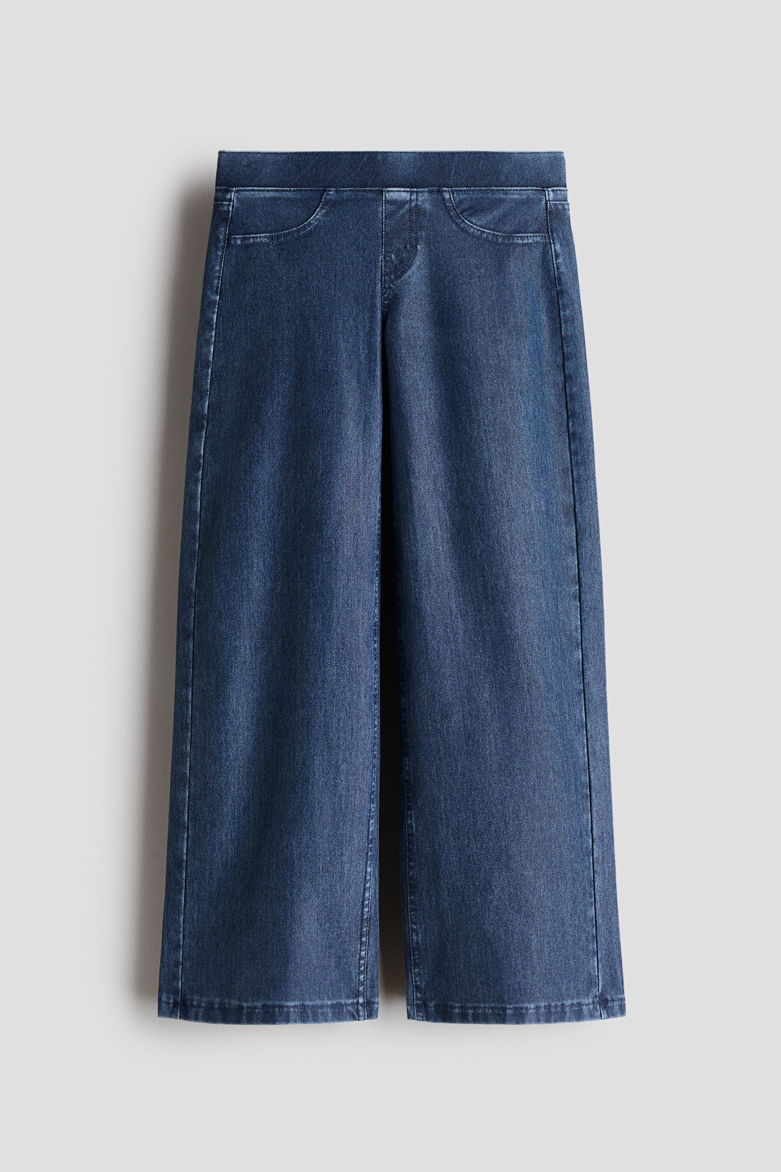 A pair of wide-leg denim trousers in a dark blue wash is hanging front-facing, featuring a flat pull-on waistband and mock curved front pockets. The garment falls into a relaxed wide-leg silhouette with subtle fading throughout the fabric.