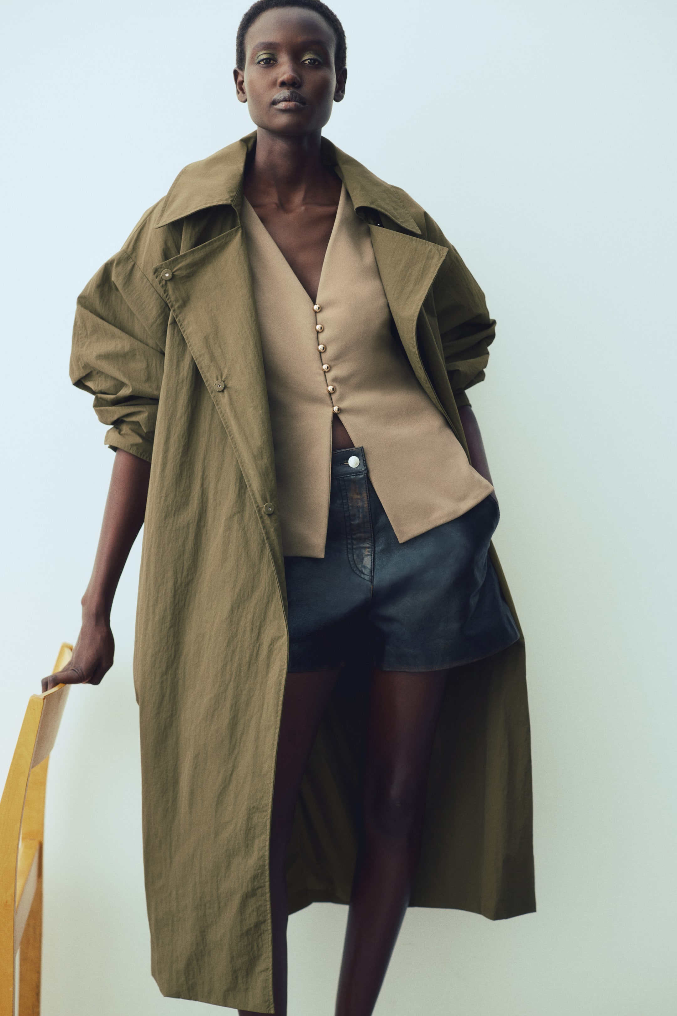 Xem ảnh lớn hơn: Chiếc áo gile màu be ôm vừa vặn, cổ chữ V với các cúc kim loại hình vòm chạy xuống tới eo, tạo độ loe nhẹ ở gấu, được người mẫu mặc. Nó được mặc bên dưới một chiếc trench coat màu xanh ô liu đang mở.