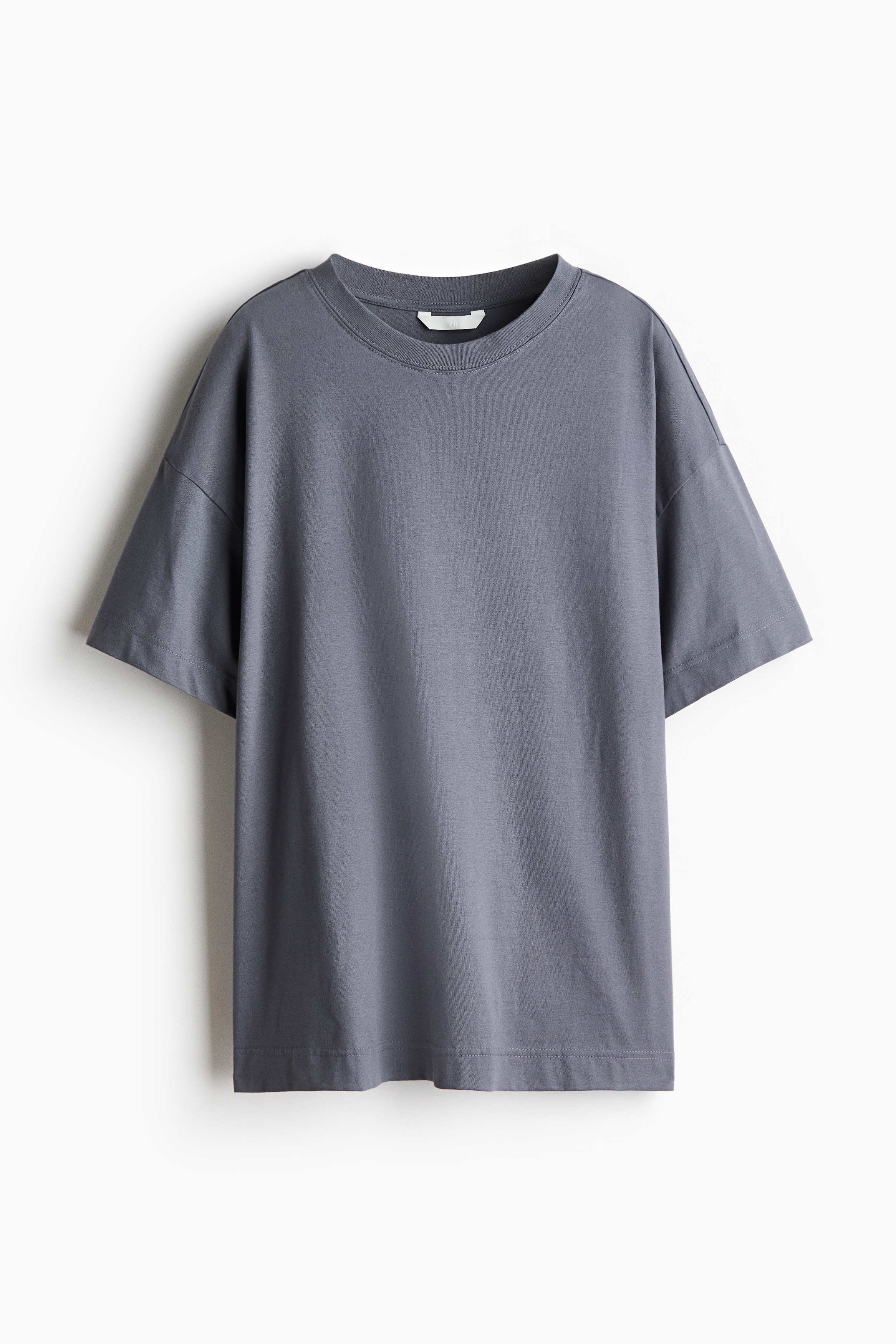 Una T-shirt oversize in jersey di cotone grigio è indossata dalla modella, sovrapposta a un top bianco a maniche lunghe. La T-shirt presenta spalle scese, un orlo dritto e un girocollo rifinito a costine, che la modella sta toccando delicatamente con una mano.