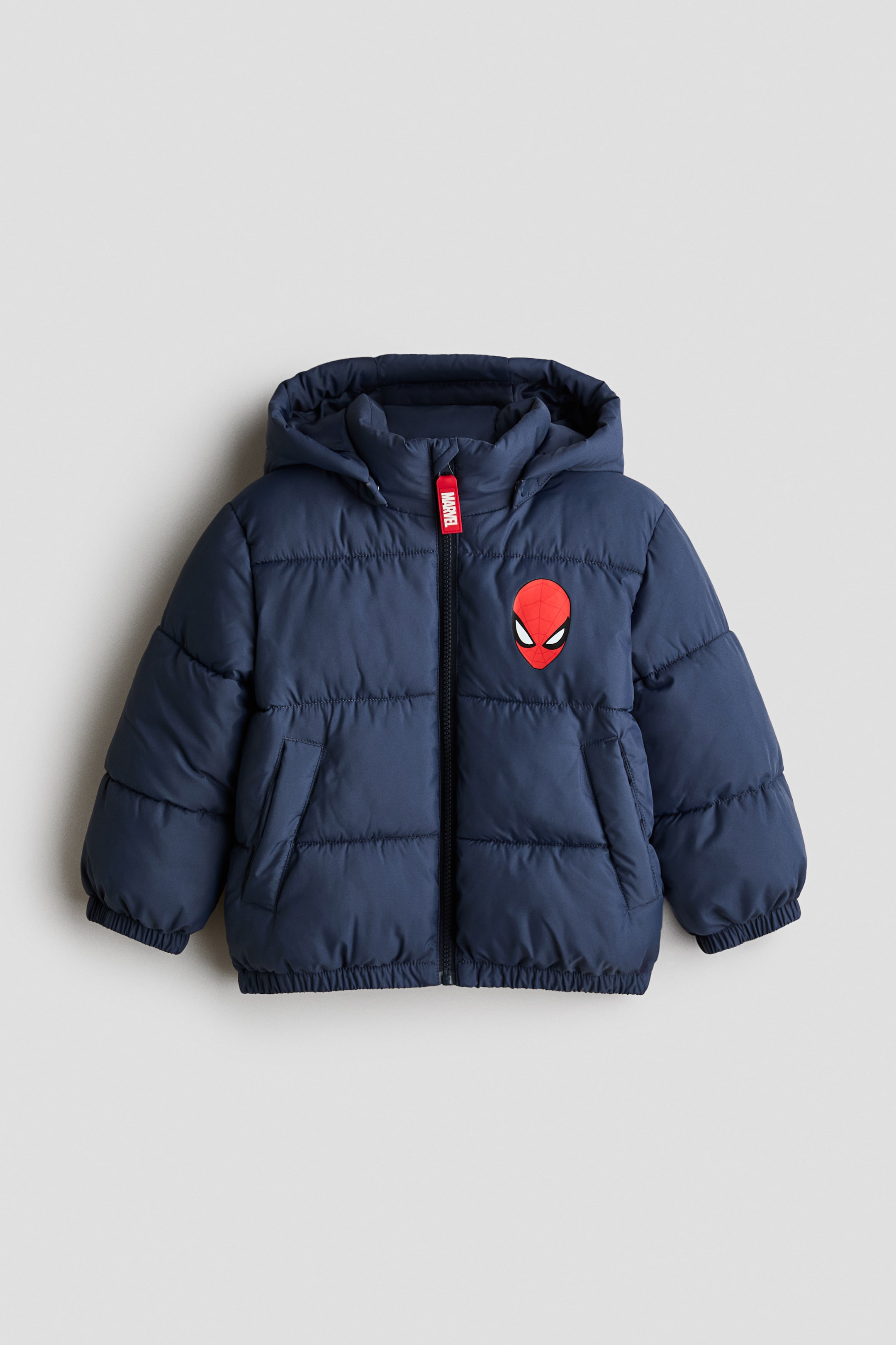 View larger image: A children’s navy blue hooded puffer jacket is shown from the front, featuring a quilted design with a central zip fastening and a red ‘MARVEL’ pull tab. The jacket features a red Spider-Man mask patch on the left chest, along with two side welt pockets and elasticated cuffs and hem.
