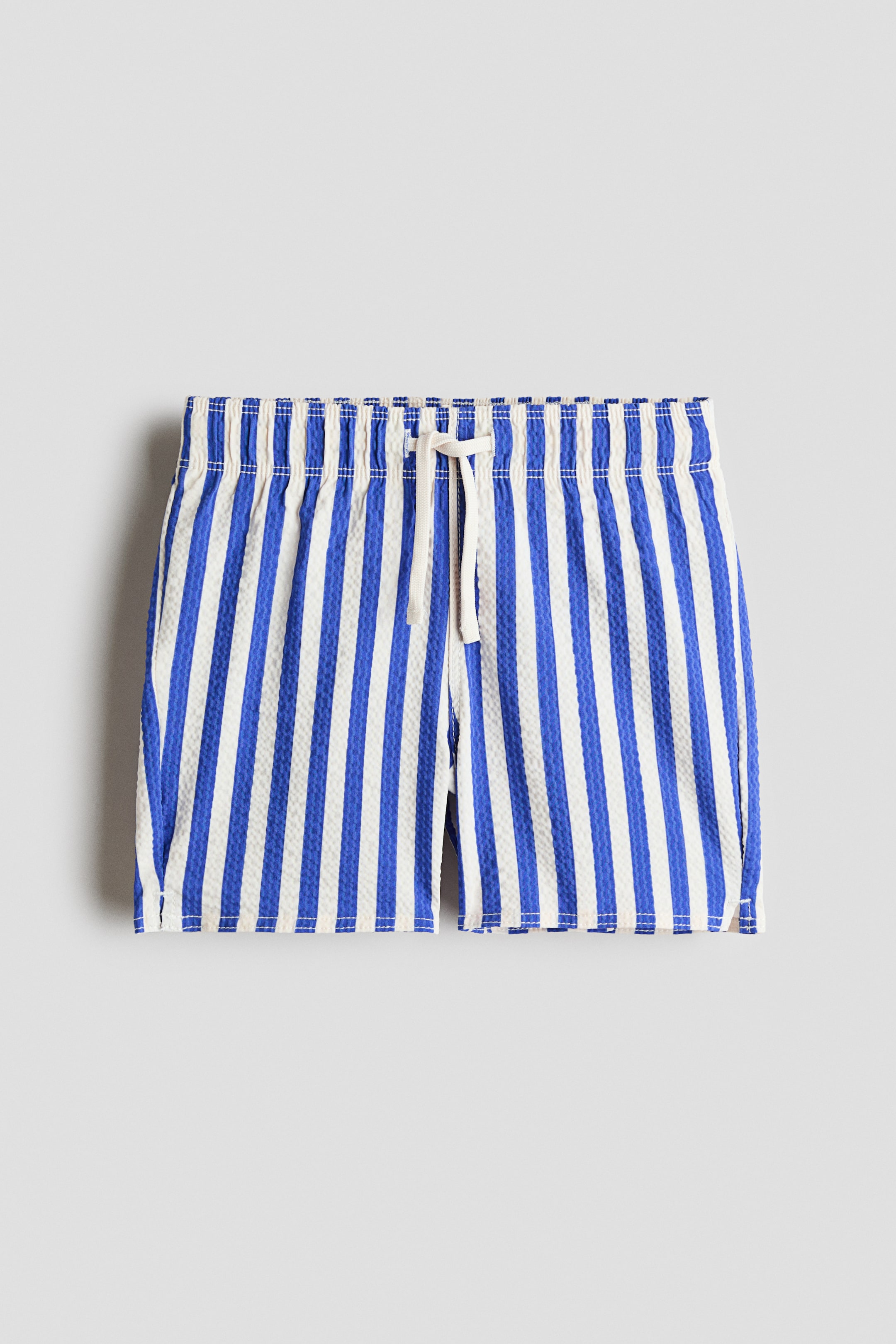 Grotere afbeelding bekijken: De H&M Adorables zwemshorts worden van voren getoond, met blauwe en witte verticale strepen op een gestructureerde stof. Ze zijn ontworpen met een elastische tailleband met trekkoord, waarbij een wit trekkoord in het midden is vastgebonden, en een kleine split zichtbaar is aan één kant van de zoom.