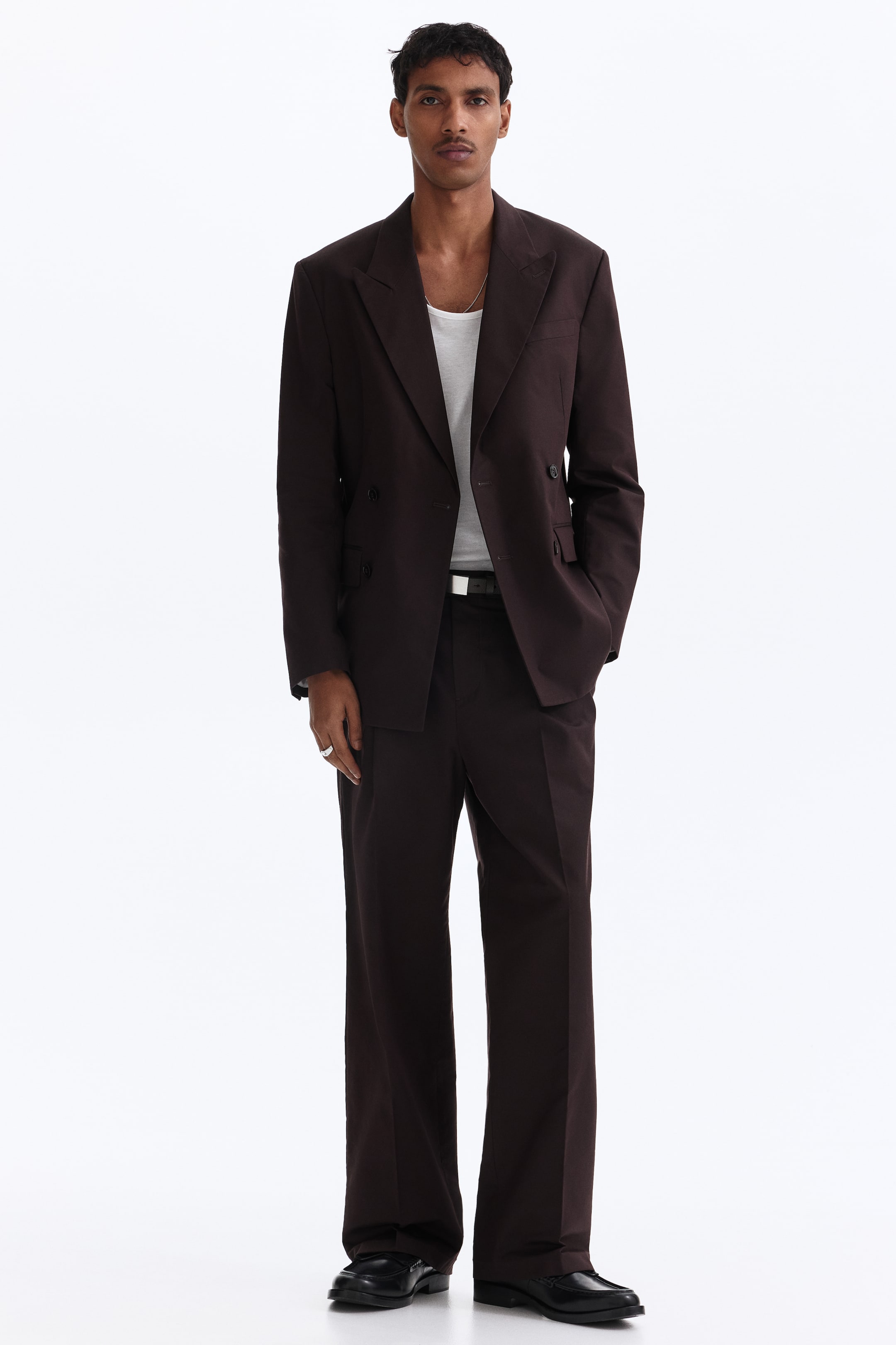 Ingrandisci l'immagine: Un completo doppiopetto marrone scuro presenta un blazer a vestibilità rilassata con ampi revers a punta e sei bottoni sul davanti, abbinato a pantaloni coordinati a gamba larga. Una canotta bianca è visibile sotto il blazer aperto e una cintura nera stringe la vita dei pantaloni.