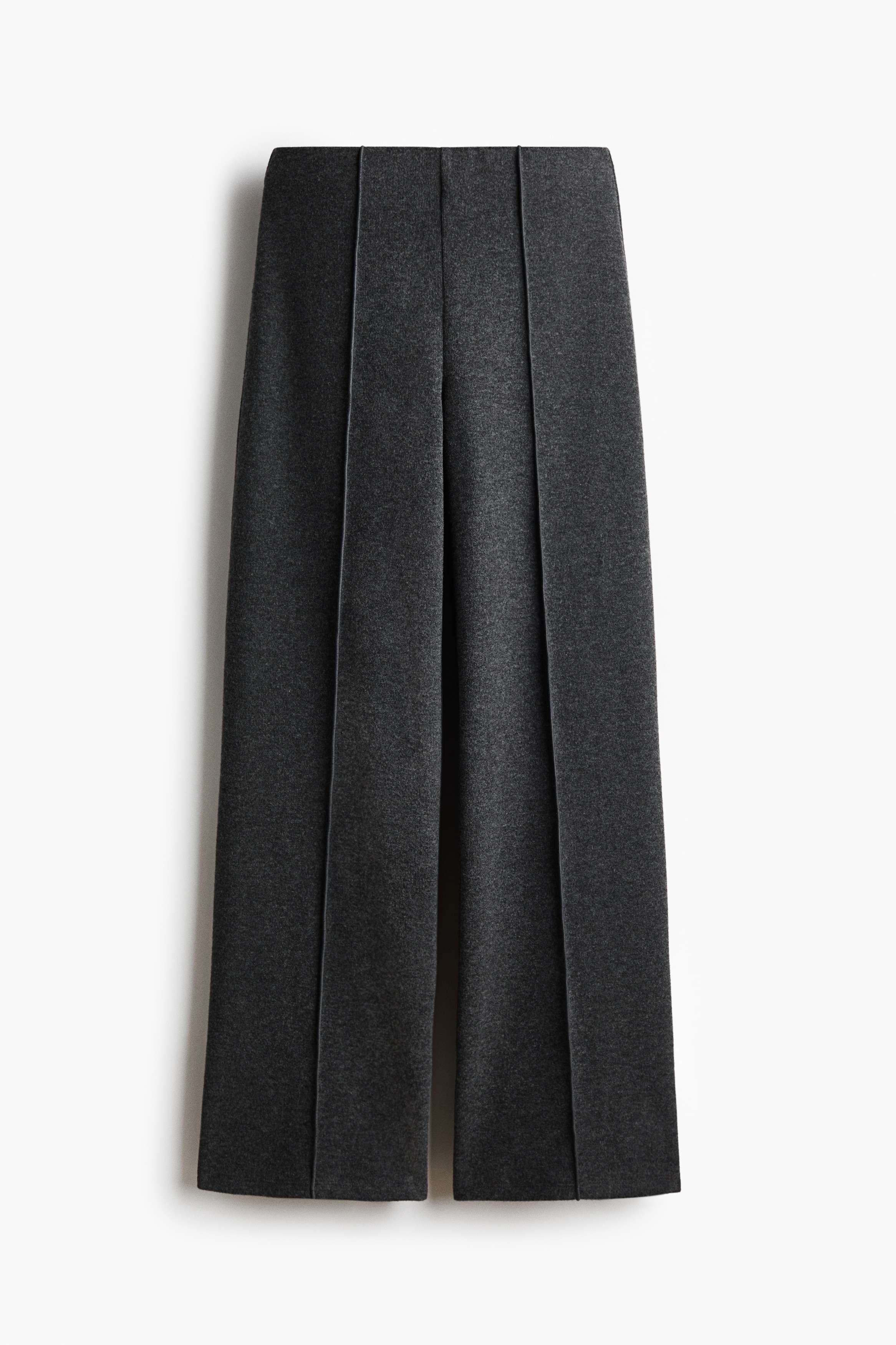 Dark grey knit trousers featuring a loose fit, straight legs, and sewn-in creases down the front are being worn by the model, who is holding a matching dark grey cardigan behind their waist. The trousers are styled with a white short-sleeved T-shirt.