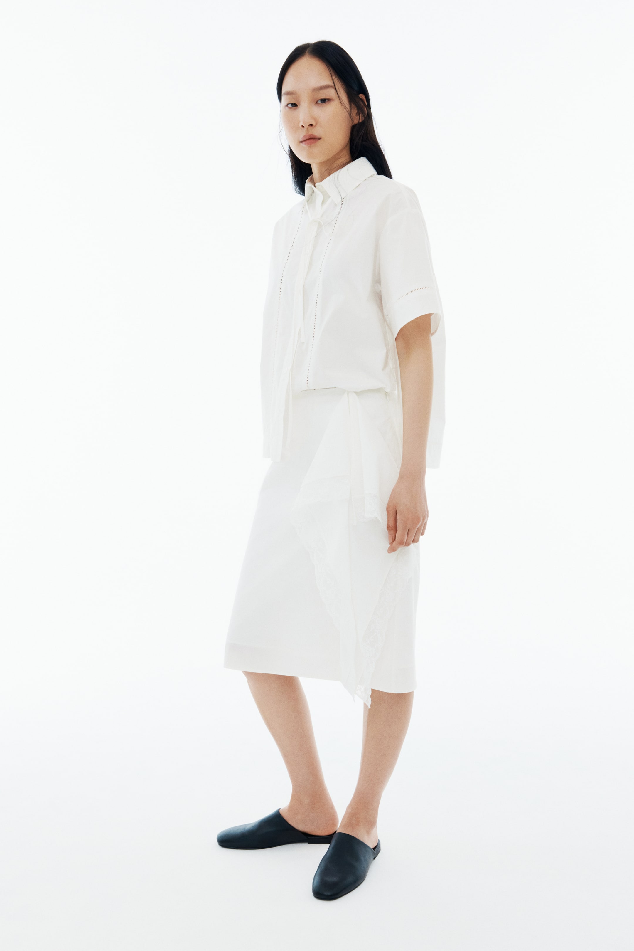 View larger image: The model is wearing a white short-sleeved collared shirt with intricate eyelet detailing on the placket and cuffs. Paired with this is a white midi skirt that boasts an asymmetric hem and delicate lace trim. Completing the look, the model is donning black flat mules.