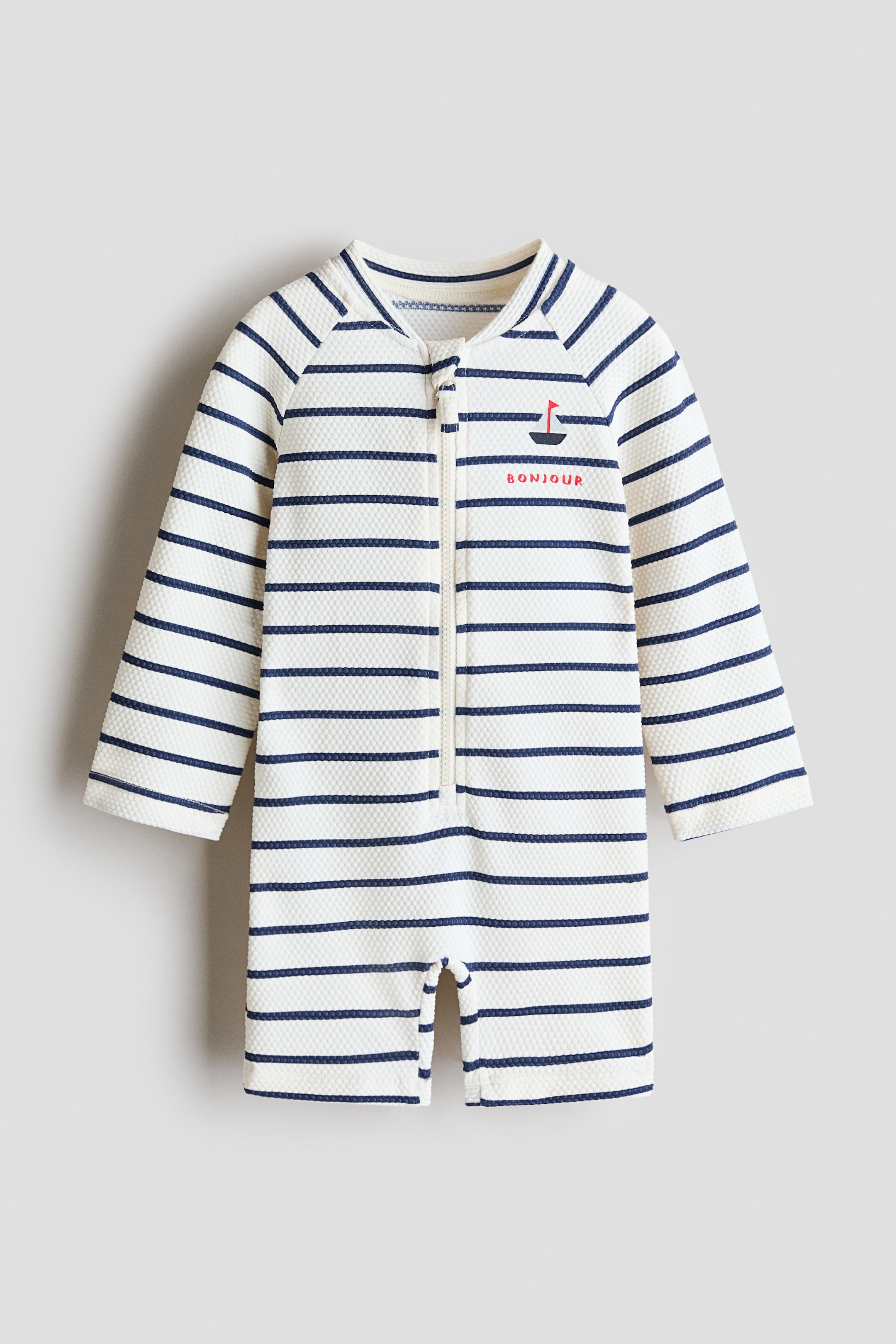 Ver imagem maior: Um fato de banho texturizado, virado para a frente, exibe riscas horizontais em creme e azul-marinho. Apresenta uma pequena gola alta, mangas compridas raglan, pernas curtas e fecho de correr frontal. Um pequeno barco à vela bordado com o texto "BONJOUR" adorna o peito esquerdo.