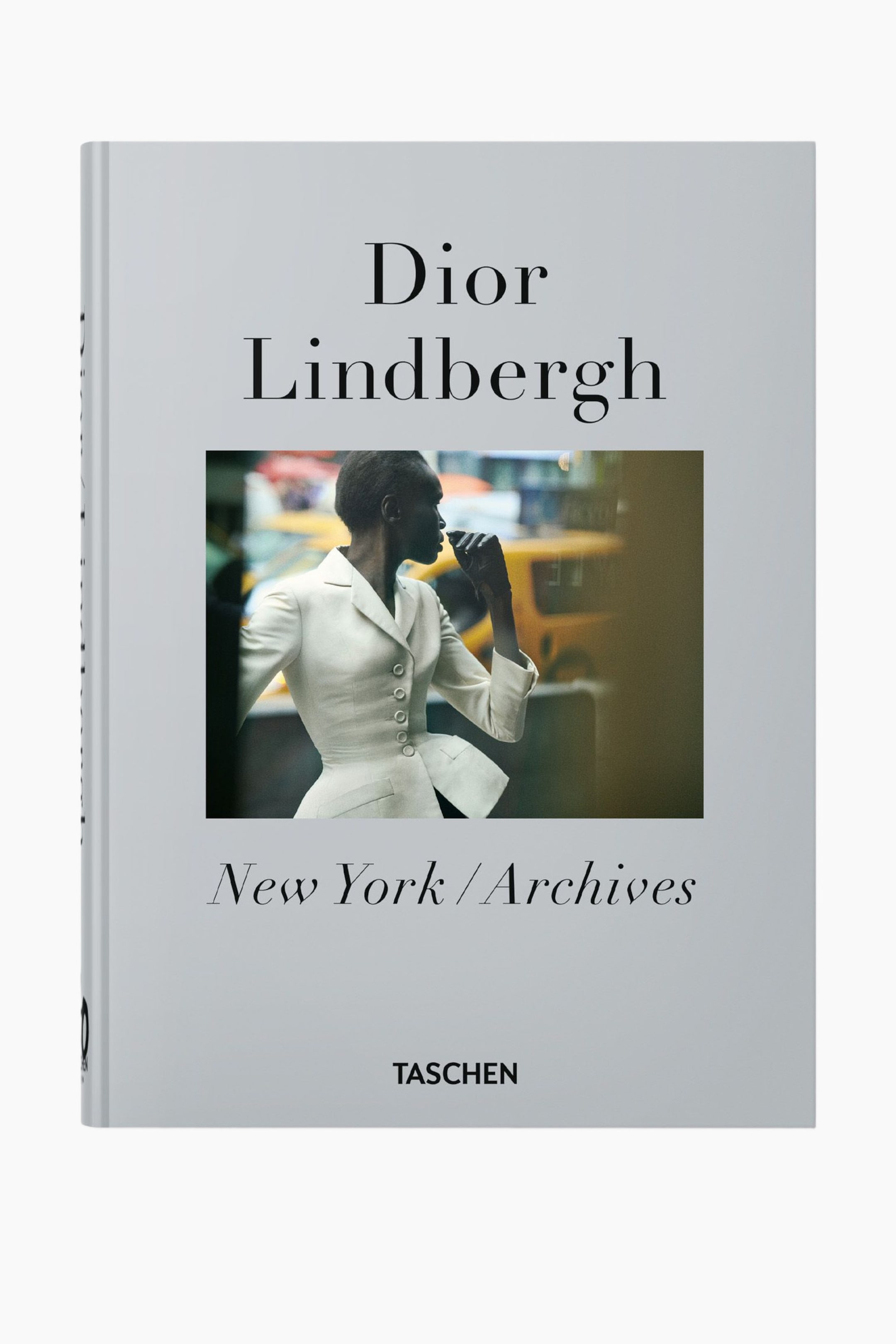 Se større billede: En bog vises med et lysegråt omslag, let vinklet så en del af ryggen er synlig. Omslaget har "Dior Lindbergh" i sort tekst, placeret over et centralt billede af en person i profil iført en hvid, figursyet jakke med fem knapper og peplum-talje. Under billedet ses "New York / Archives" og "TASCHEN" i sort tekst.